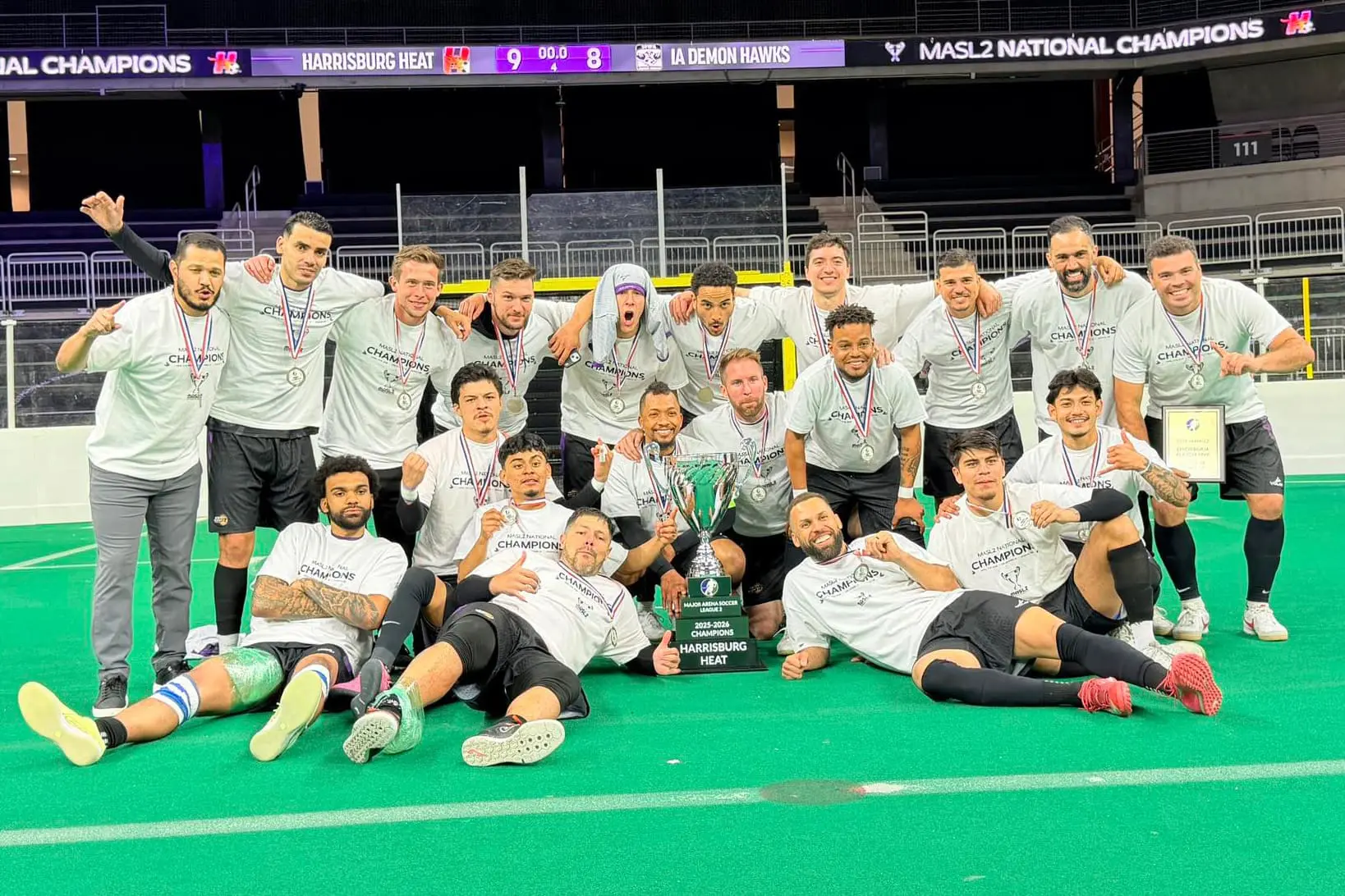 Group of male soccer players in gray shirts pose on a green turf with medals, a large trophy, and a plaque after winning a championship.