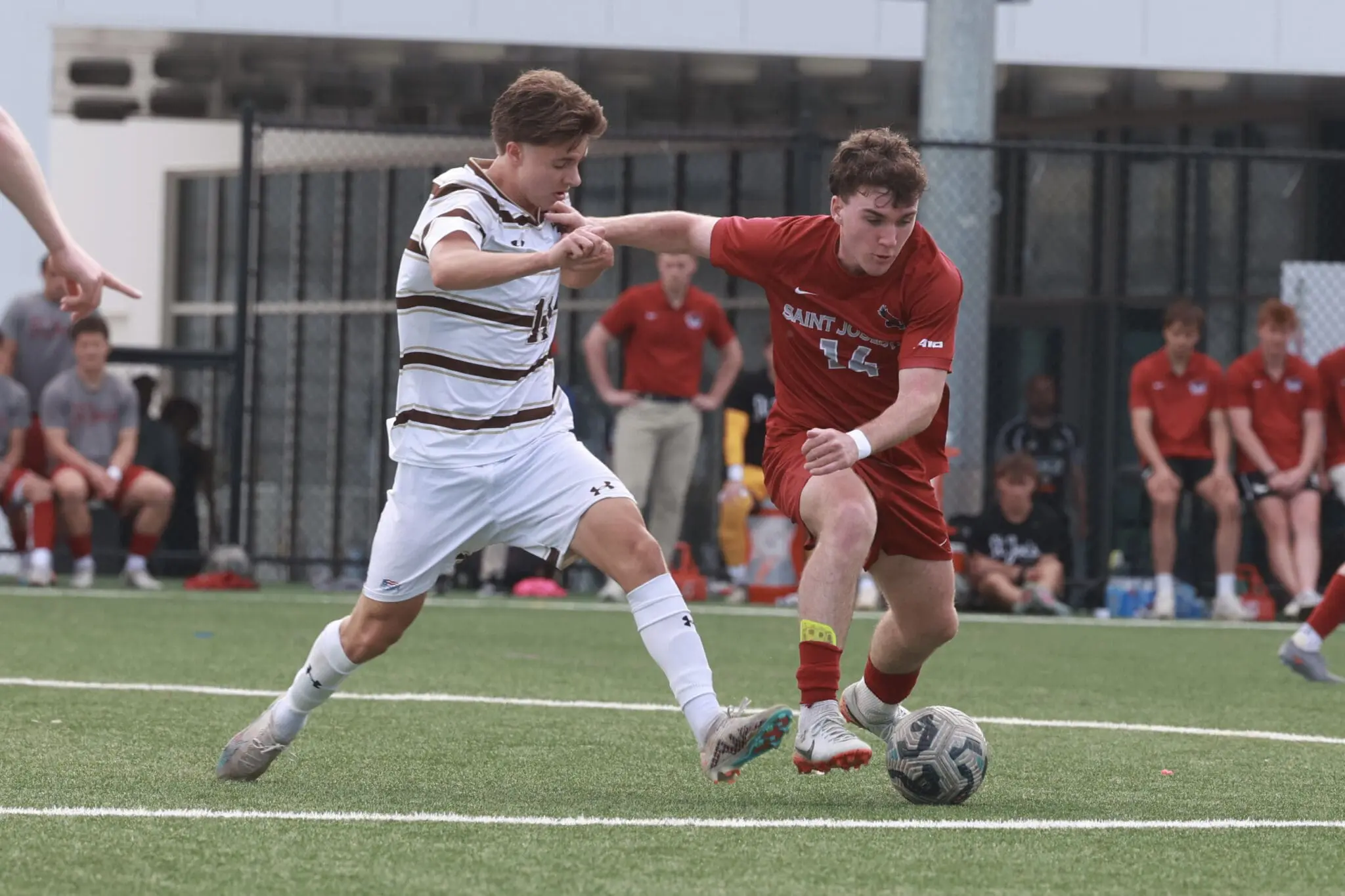 Soccer players in red and white stripes duel for the ball on a turf field, with onlookers on the sideline in the background.
