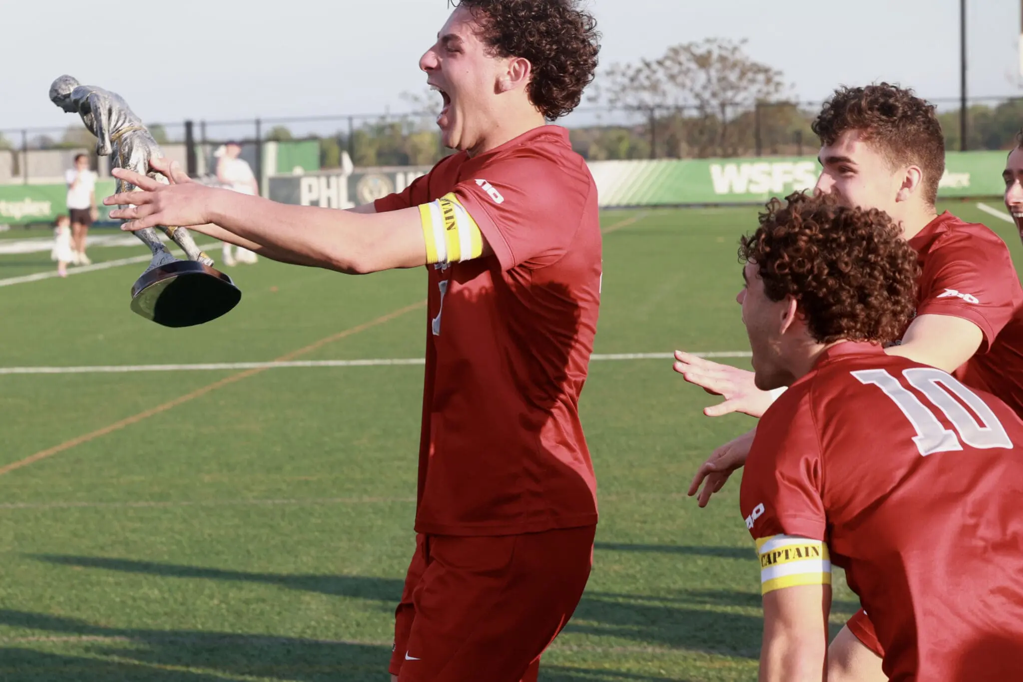 Soccer players in red celebrate on a green field as one lifts a silver trophy in triumph.