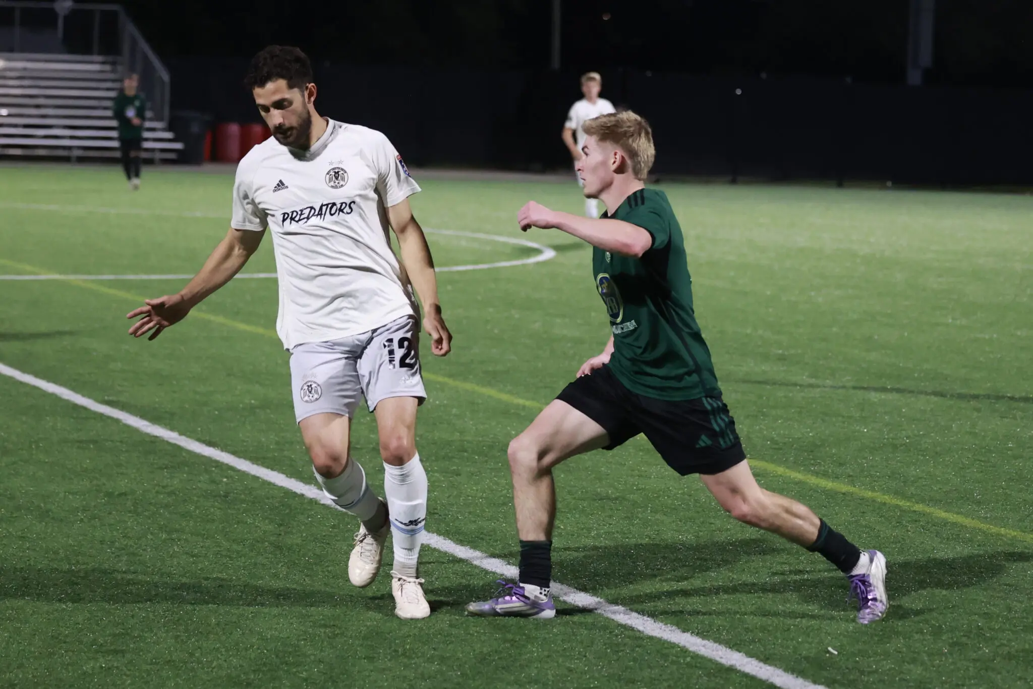 Two soccer players race for the ball on a night game; white team with 'Predators' kit vs a green opponent on a green turf field.