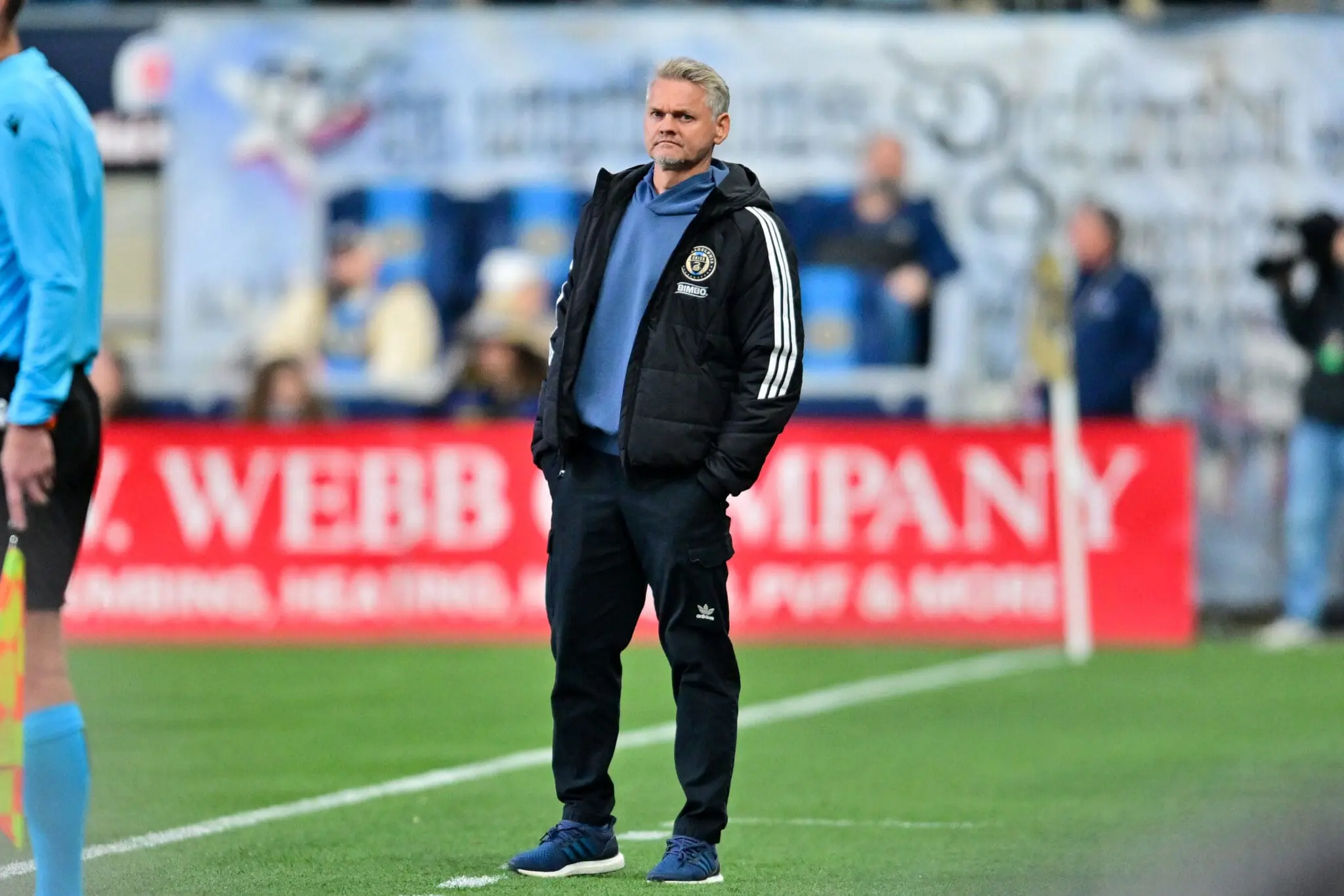 Soccer coach standing on the sideline in a black Adidas jacket with white stripes, blue hoodie underneath, watching the game.