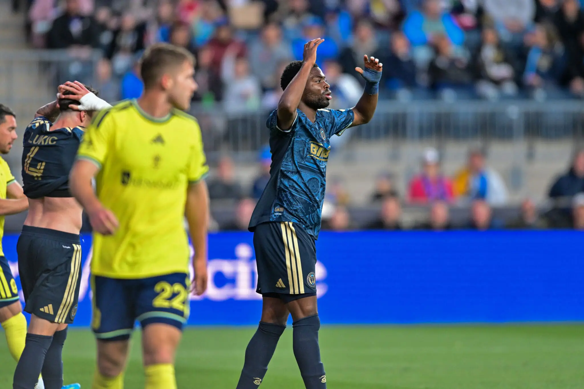 Soccer player in blue raises arms in celebration while teammates in yellow and blue look on on a green field with a blurred crowd in the background.