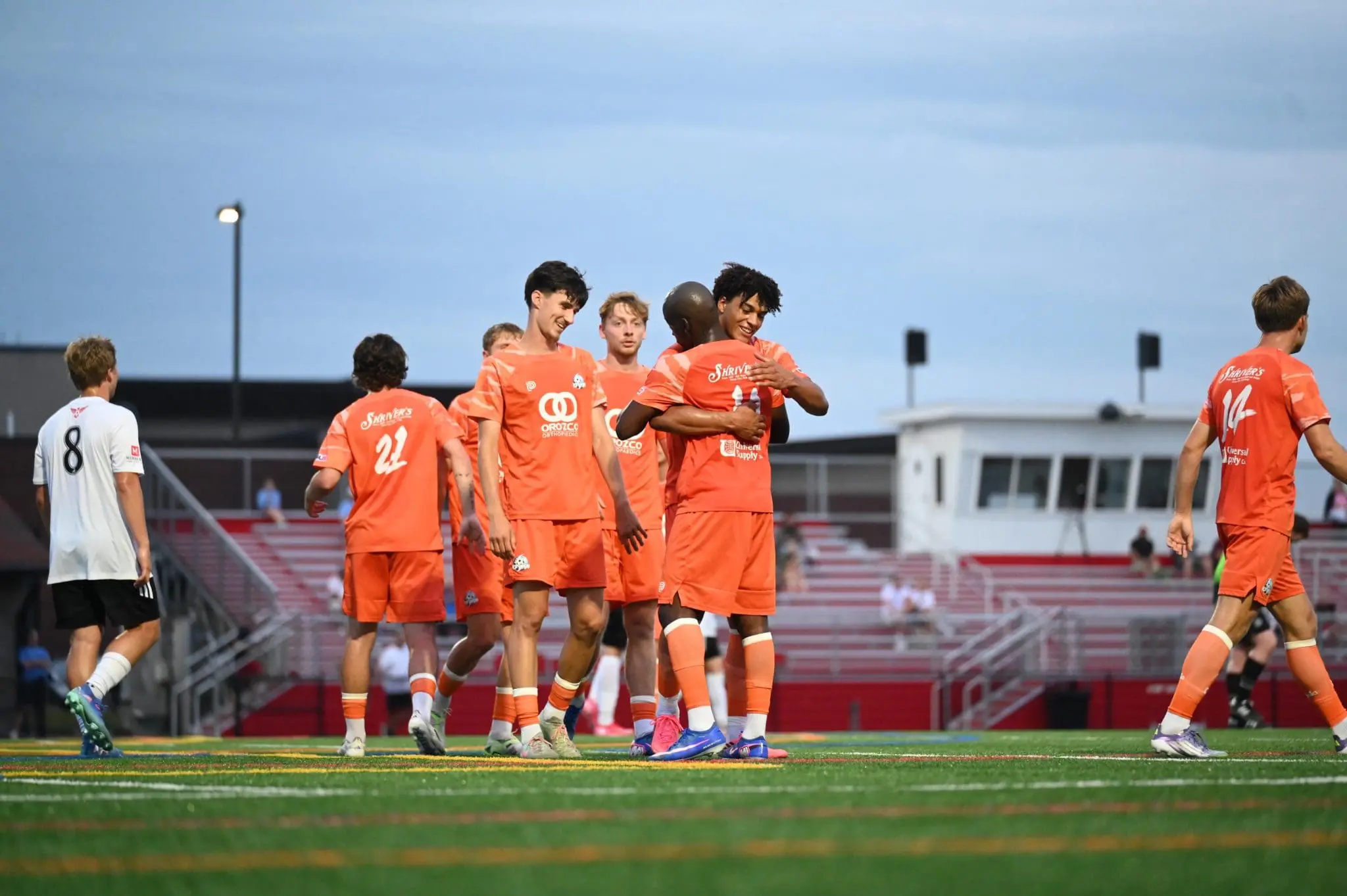 Soccer players in orange jerseys celebrate together on the field, embracing near the goal area with a white-uniform opponent nearby.