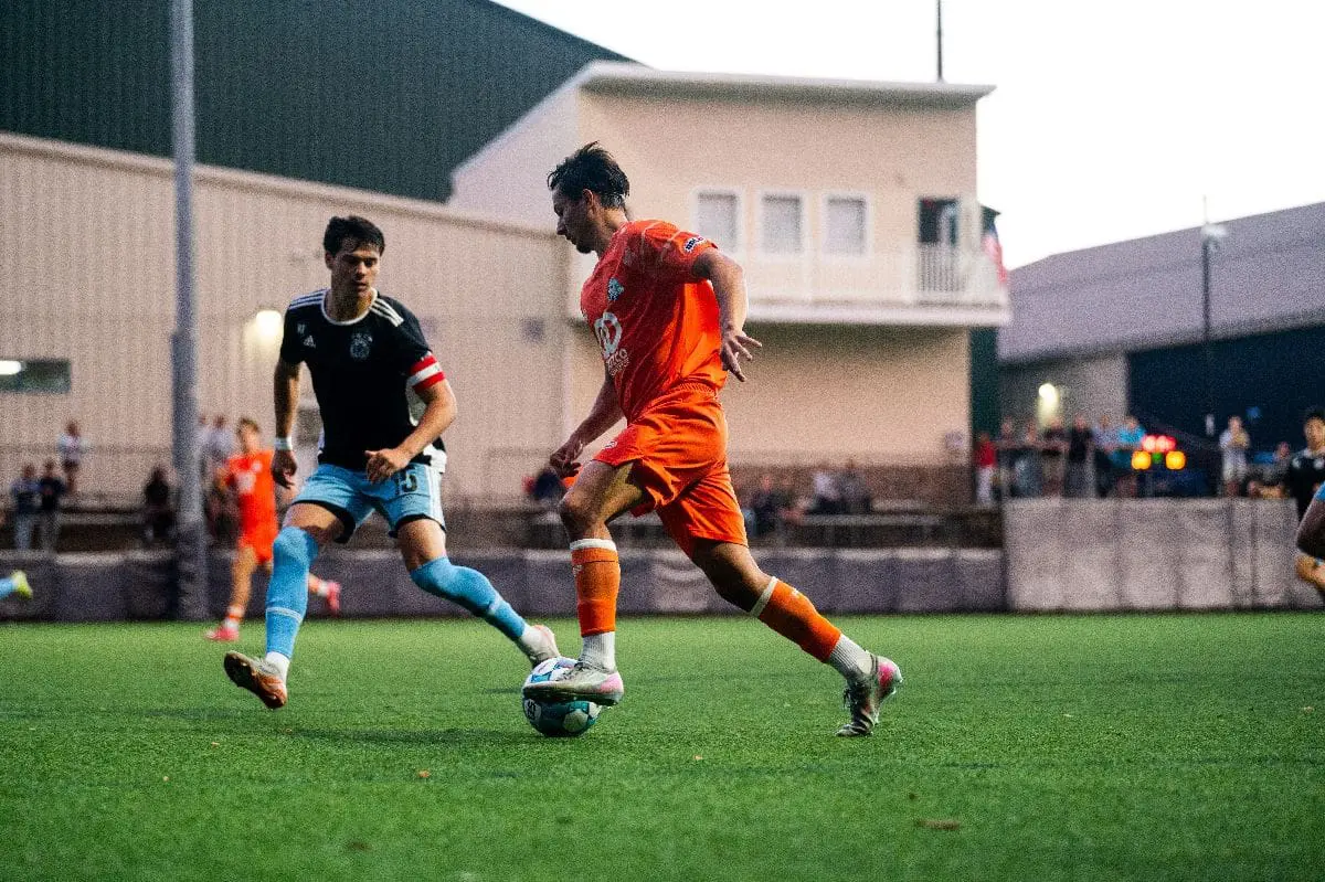 Soccer player in an orange uniform dribbles the ball past a defender in blue on a turf field.