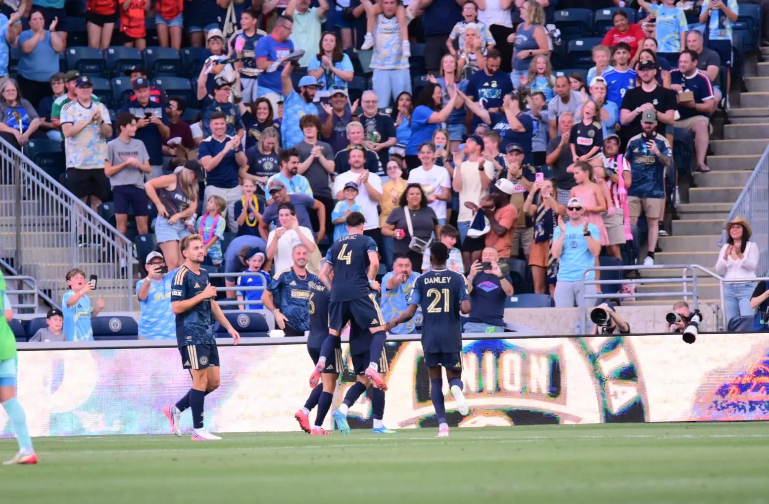 Soccer players in dark blue uniforms celebrate a goal on the field while a cheering crowd watches from the stands.