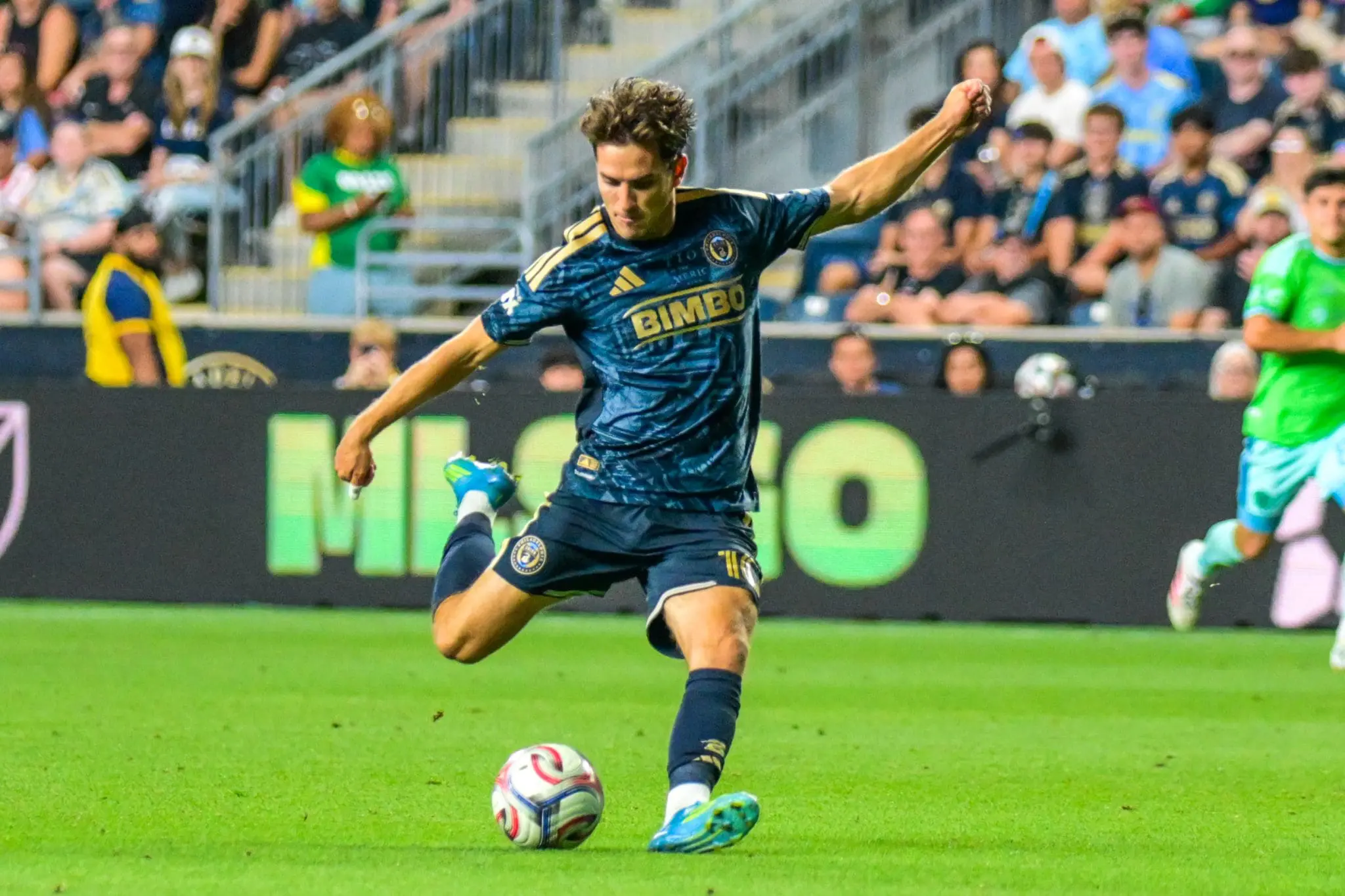 Soccer player in a blue kit mid-kick on a green pitch with cheering spectators in the stands behind him.