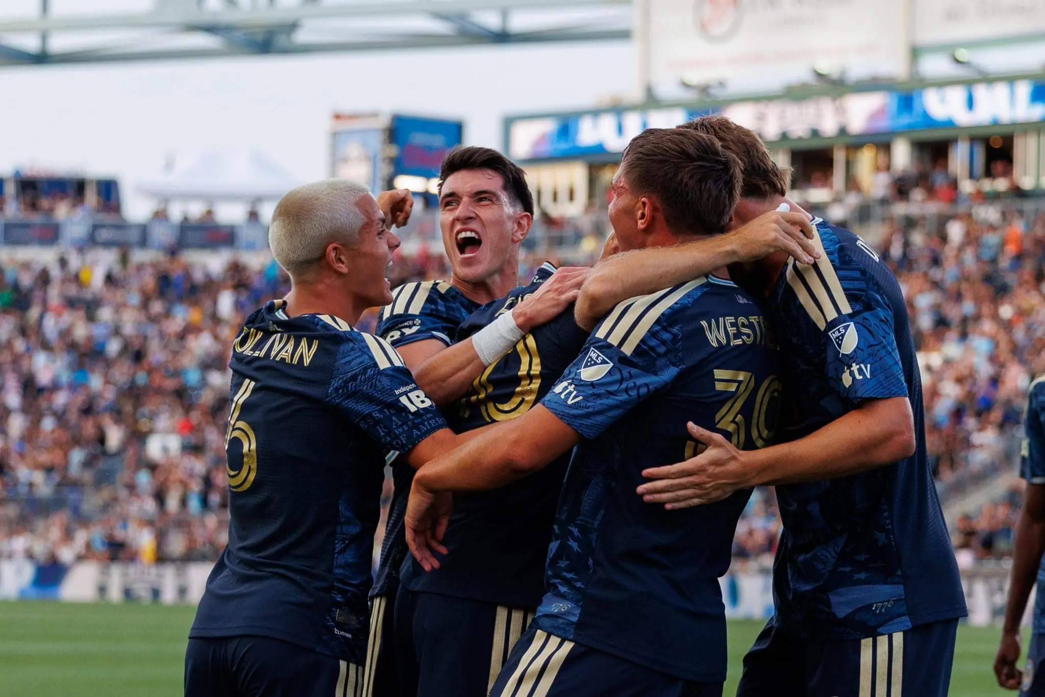 Soccer players in blue uniforms celebrate in a huddle on the field, arms around each other in a stadium full of cheering fans