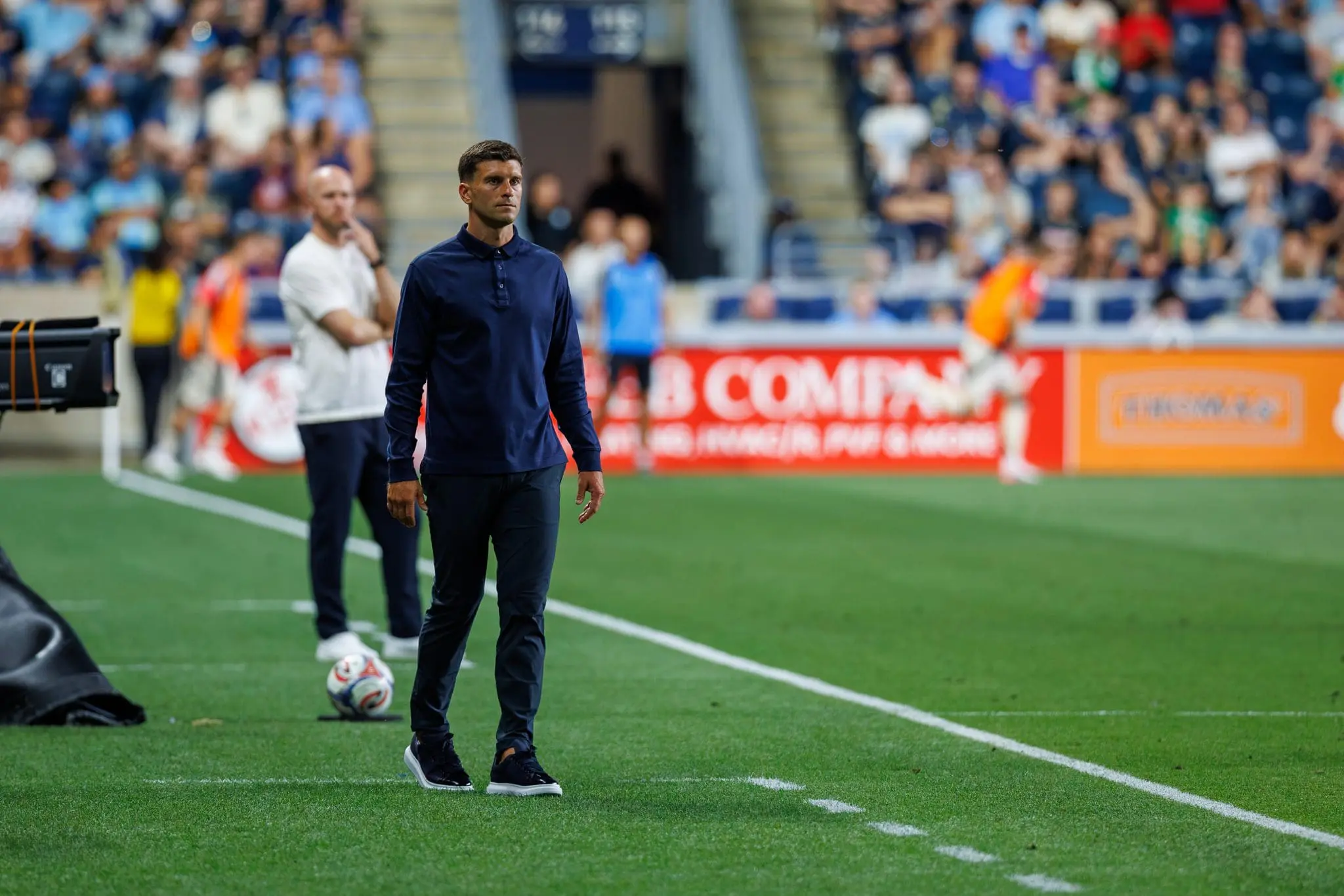 Man in a dark blue athletic outfit walks along the sideline on a soccer field, with blurred spectators in the stands behind him.