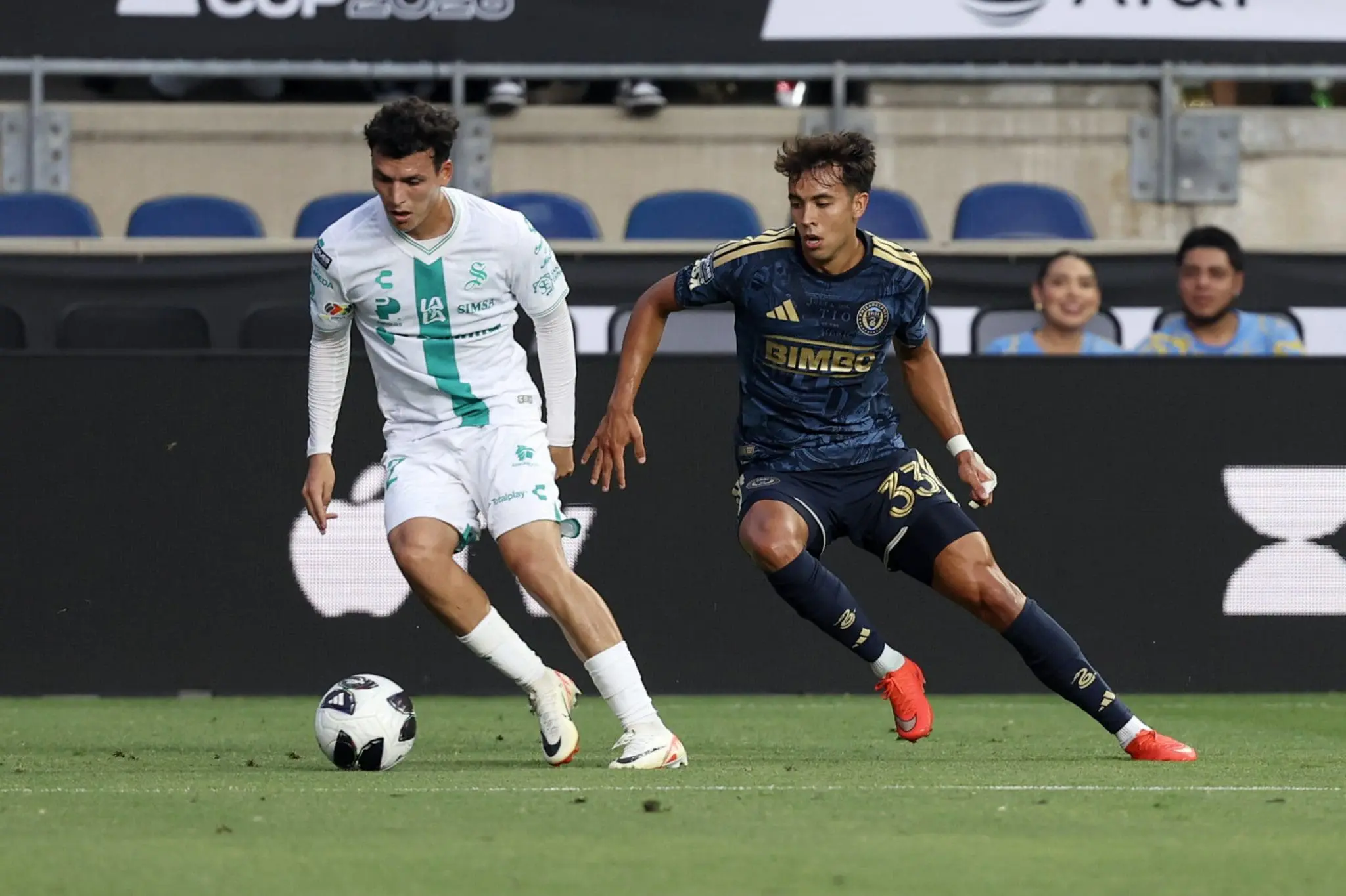 Two soccer players in white and dark blue jerseys chase the ball on a green pitch, with empty stadium seats in the background.
