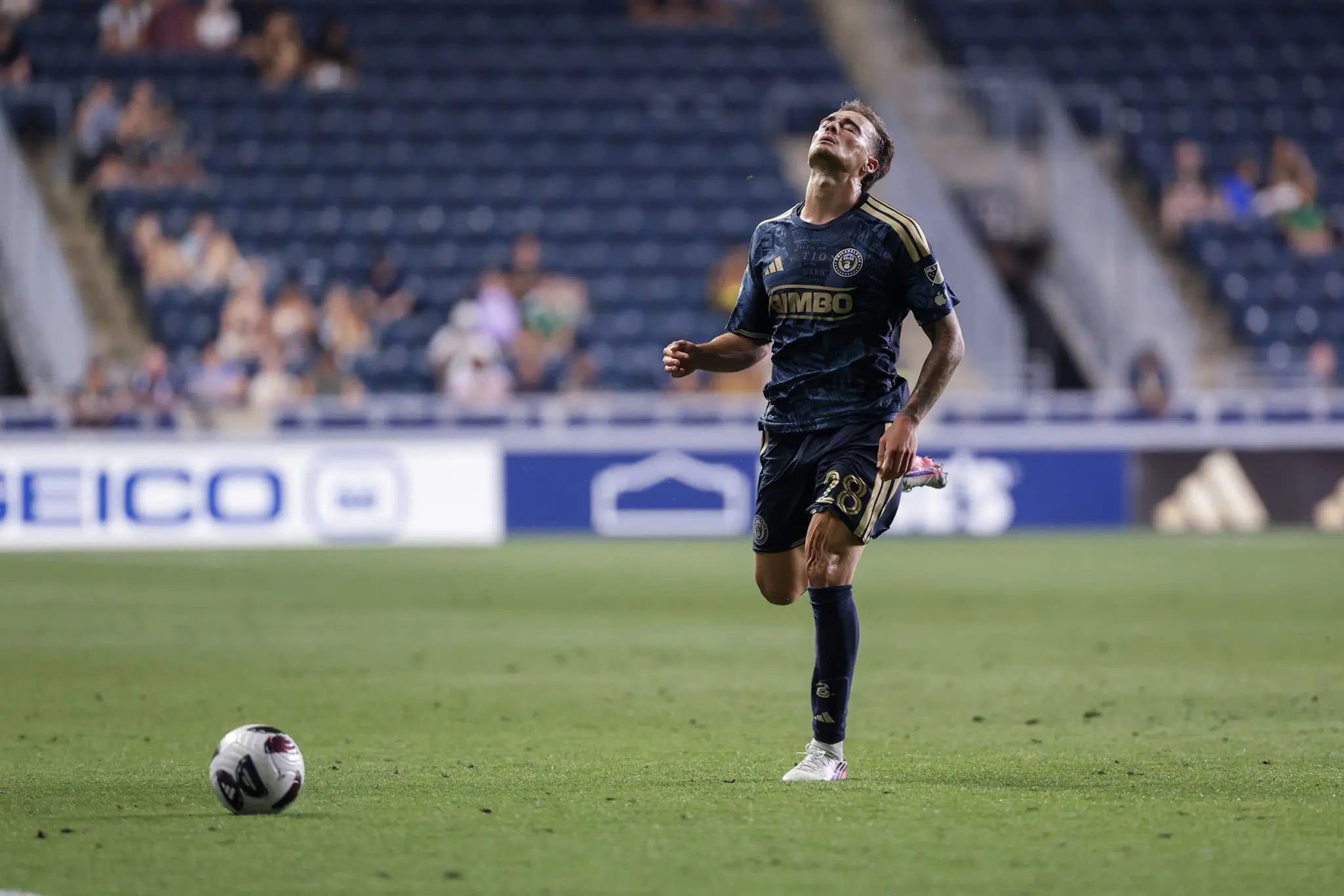 Soccer player in a dark blue jersey with gold accents runs toward a ball on a green field, stadium seats blurred in the background, number 28 on shorts visible.