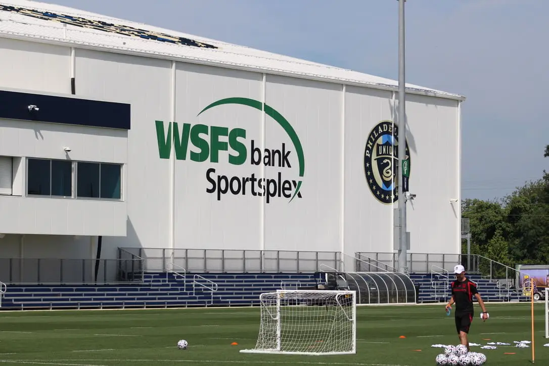 Soccer practice field in front of a large sports complex with the WSFS Bank Sportsplex sign on the building and a player near a small goal.