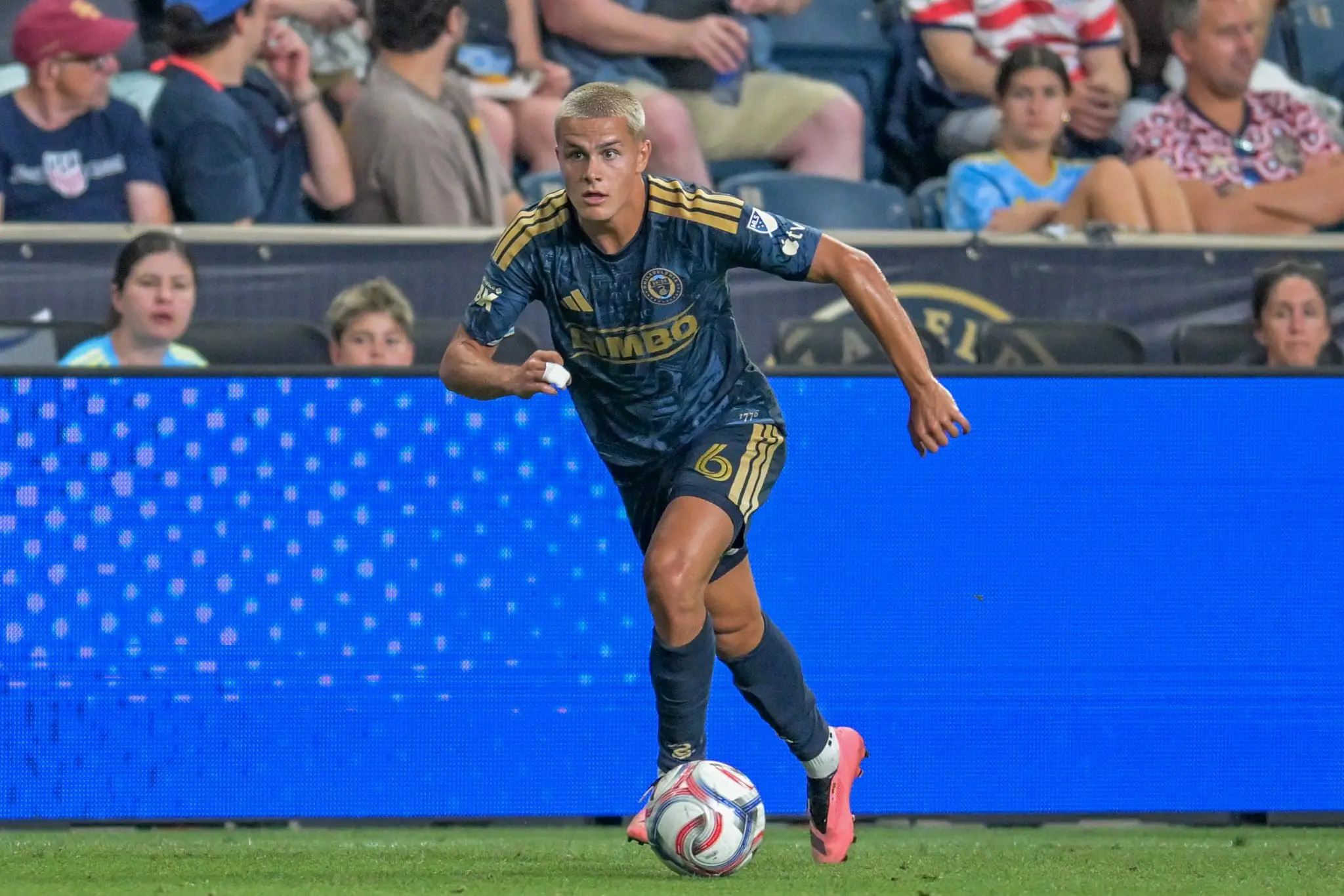 Soccer player in dark blue kit dribbles the ball on the field, with spectators in stands behind him.