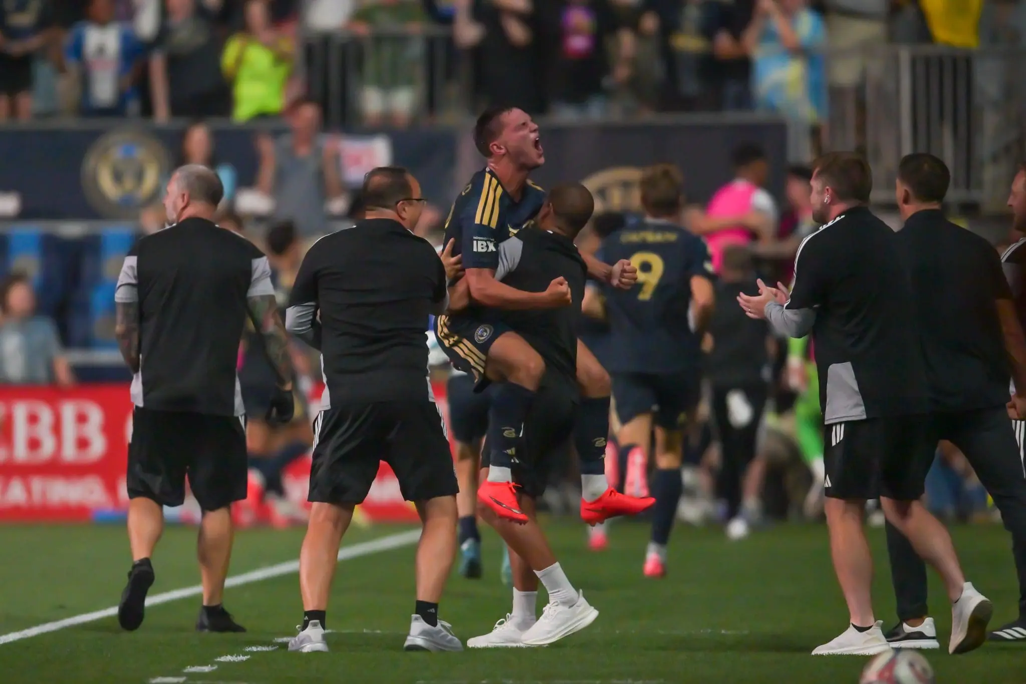 Soccer players celebrating on a green field, mid-jump with clenched fists as teammates embrace and crowd watches in stadium stands behind them