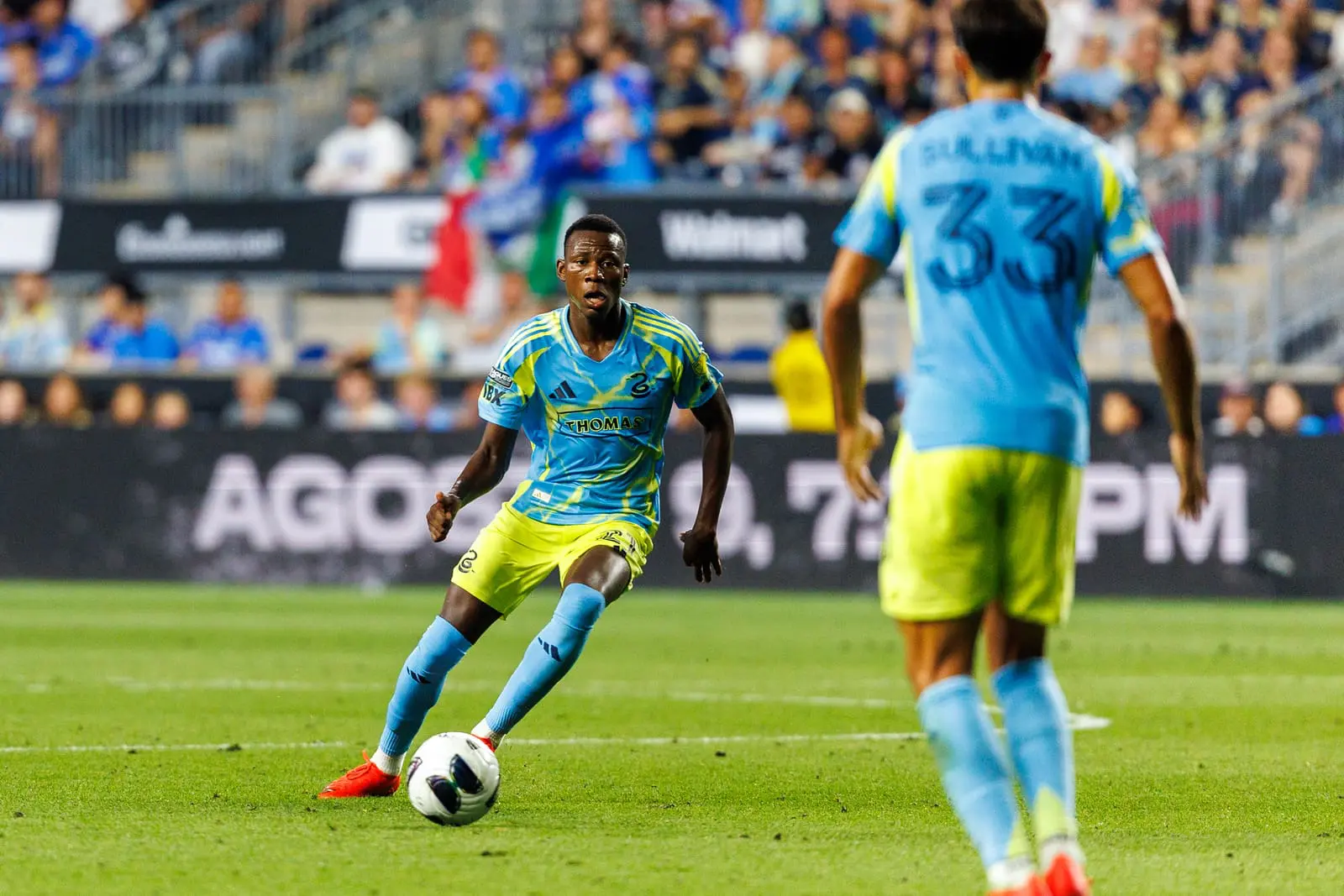 Soccer player in a sky-blue kit dribbles the ball on a green field while a teammate approaches in the foreground and spectators watch from the stands.