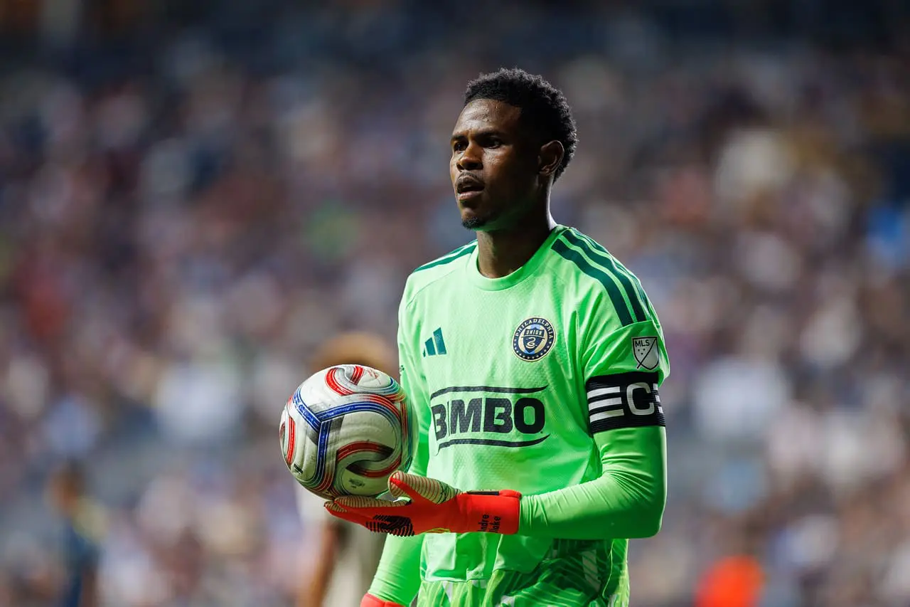 Goalkeeper in a bright green jersey holding a soccer ball on the field with a blurred crowd behind him.