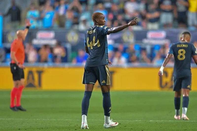 Soccer player in dark blue kit with number 14 points toward the stands while another player in back wears number 8 on a green field stadium crowd in the background