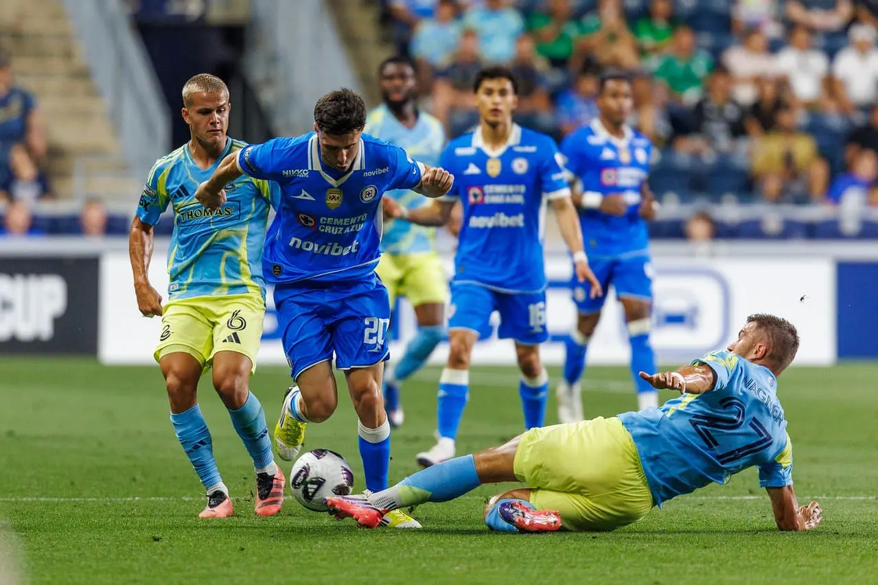 Soccer match action as blue team players contest the ball while a goalkeeper slides in to intercept on the right-hand side of the field.