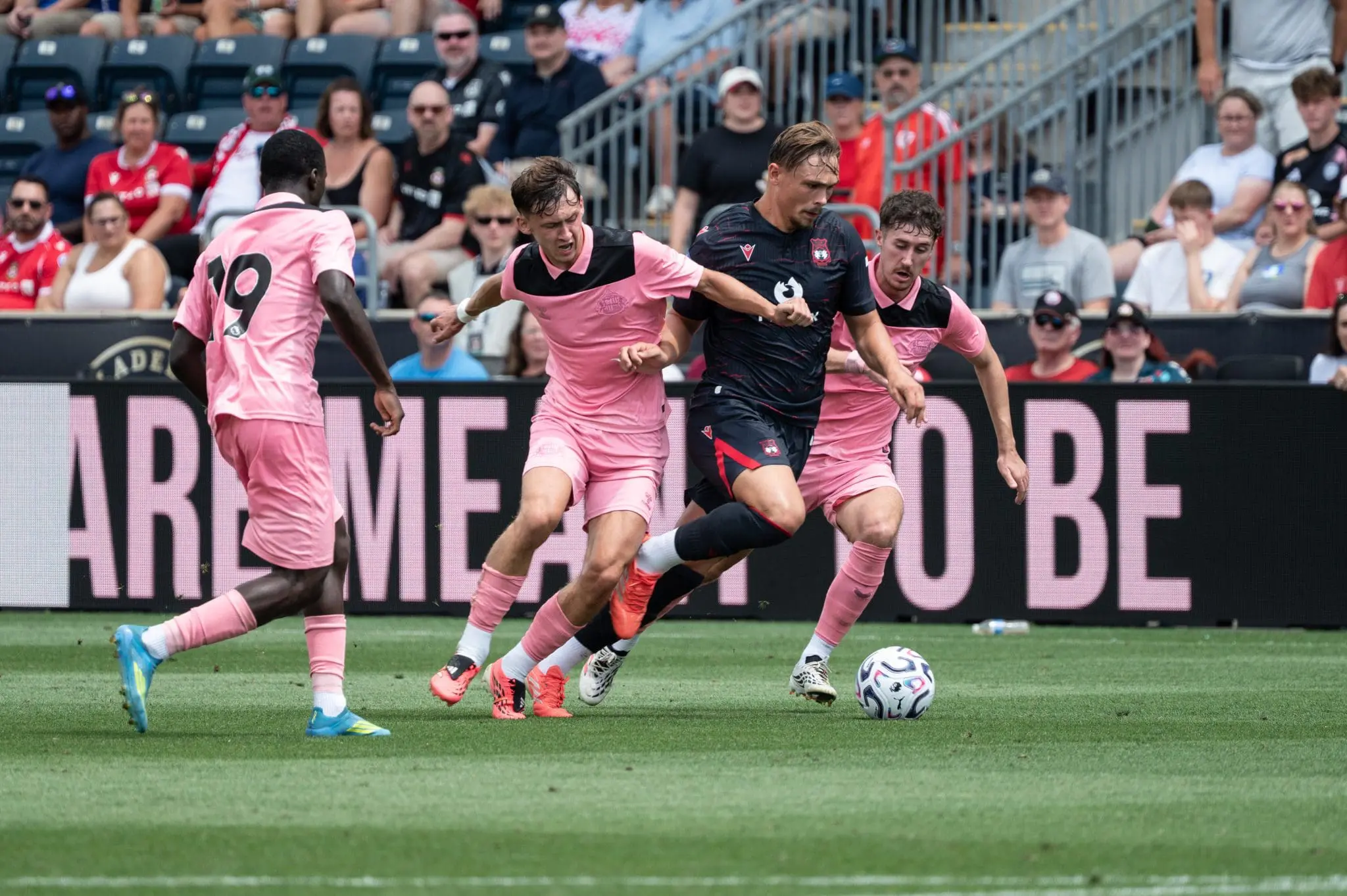 Soccer players in pink and black uniforms chase a ball on the field with fans watching from the stands in the background.