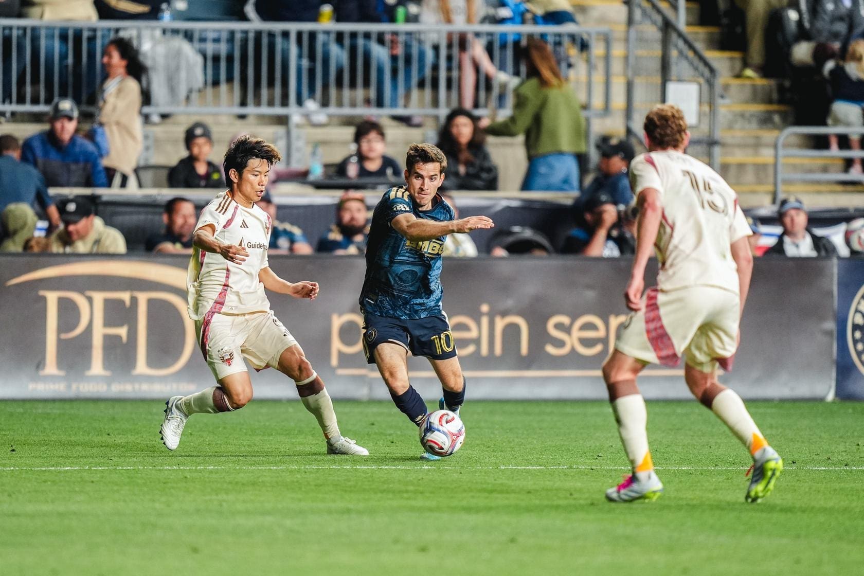 Soccer player in blue dribbles past two white-uniform opponents on a green field as spectators watch from the stands.