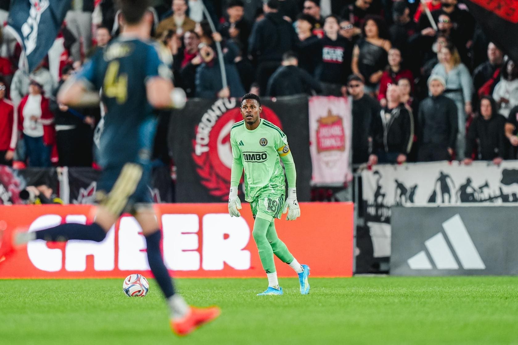 Soccer goalkeeper in a neon-green kit standing on the pitch with a ball nearby, crowd watching behind him.