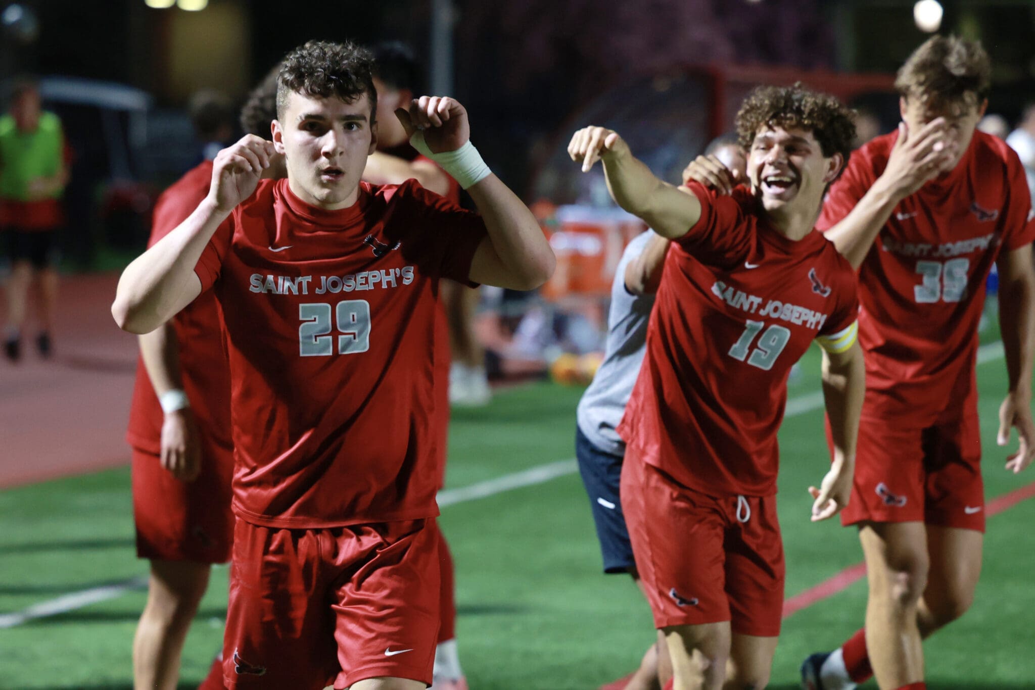 Saint Joseph's soccer players in red uniforms celebrate on the field, arms raised in victory gesture.