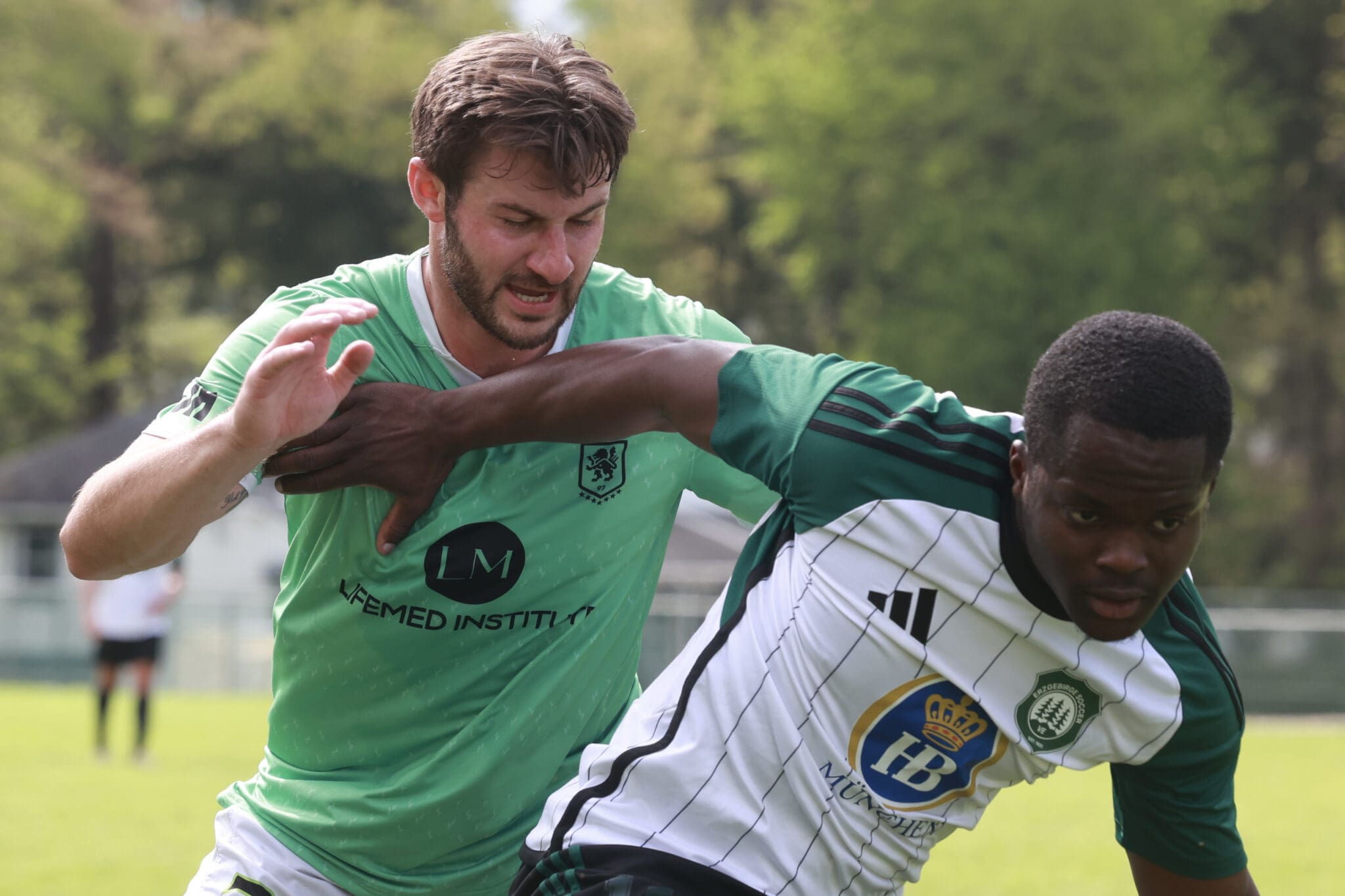 Two soccer players in green and white jerseys battle for the ball during a daytime match on a grassy field.