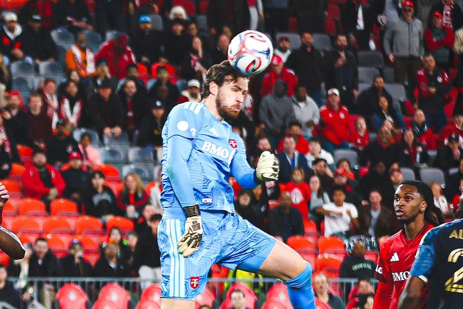 Soccer goalkeeper in a light blue kit jumps to head the ball during a match with spectators in the stands behind him.