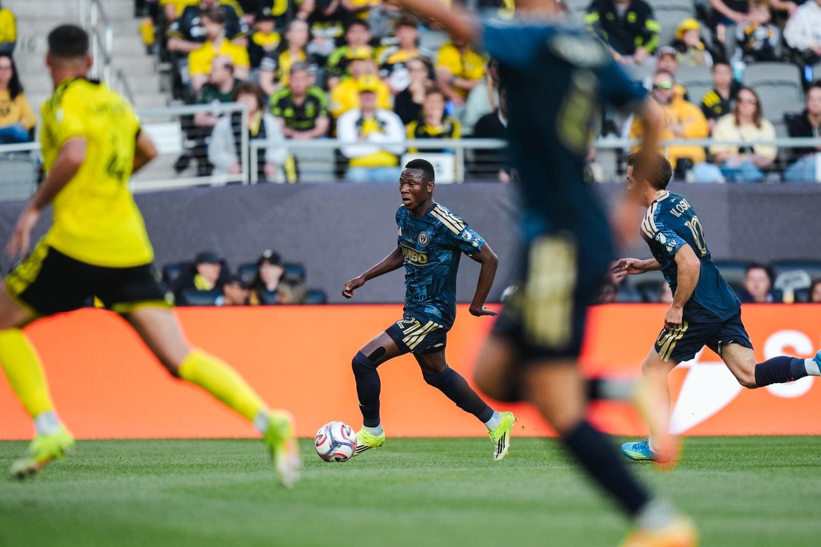 Soccer player in blue controls the ball while running forward, with yellow-clad opponents and a cheering crowd in the background.