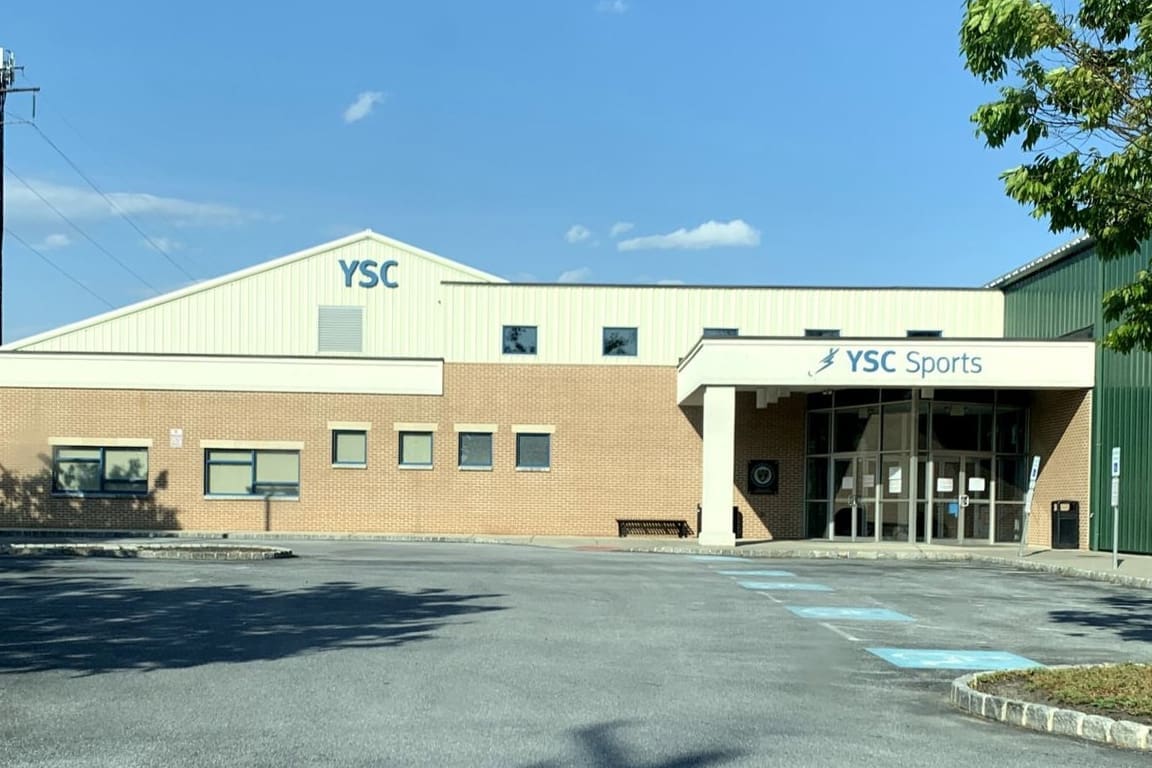 FRONT of a YSC Sports building with brick lower level and light-colored upper facade, clear glass entrance doors, and a parking lot in front
