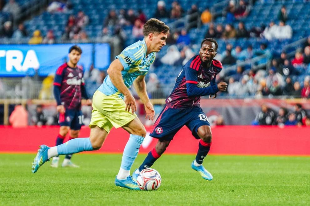 Soccer players in light blue and navy-red kits chase the ball on a green field during a match with spectators in the stands behind them.