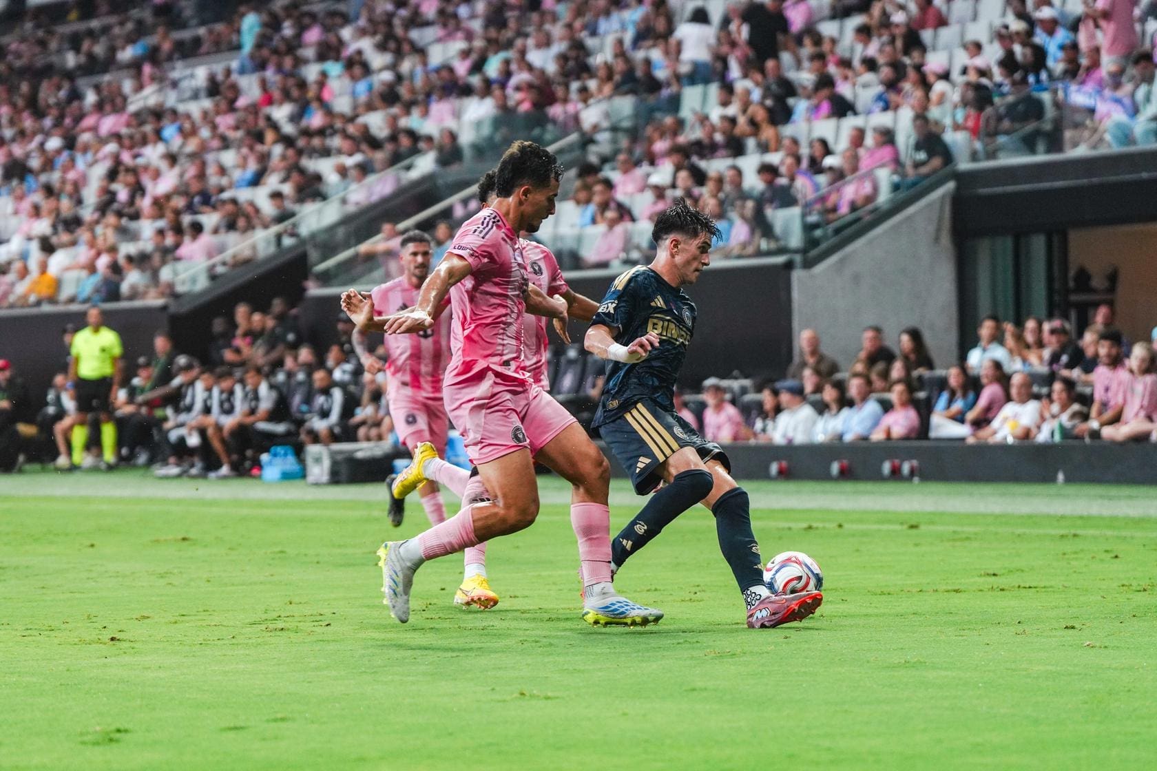 Two soccer players in pink and navy battle for the ball on a green pitch with a full stadium crowd behind them, during a match.