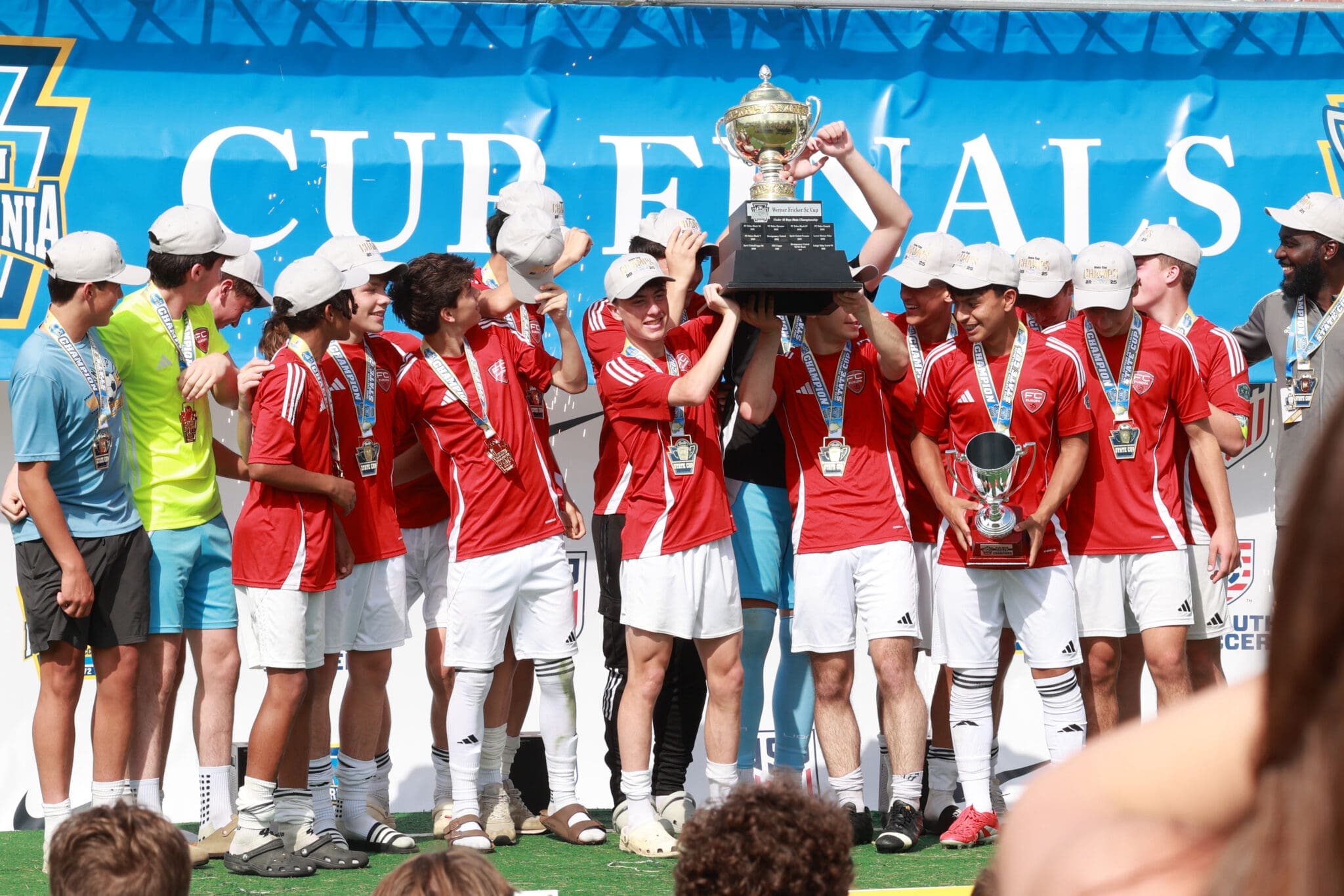 Youth soccer team celebrates victory, holding a large trophy on a podium amid spectators and medals.