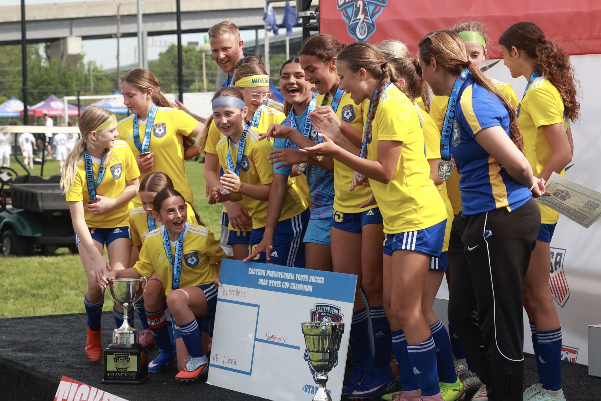 Group of young female soccer players in yellow and blue uniforms celebrate with medals and trophies at an award ceremony outdoors, smiling and posing for photos.