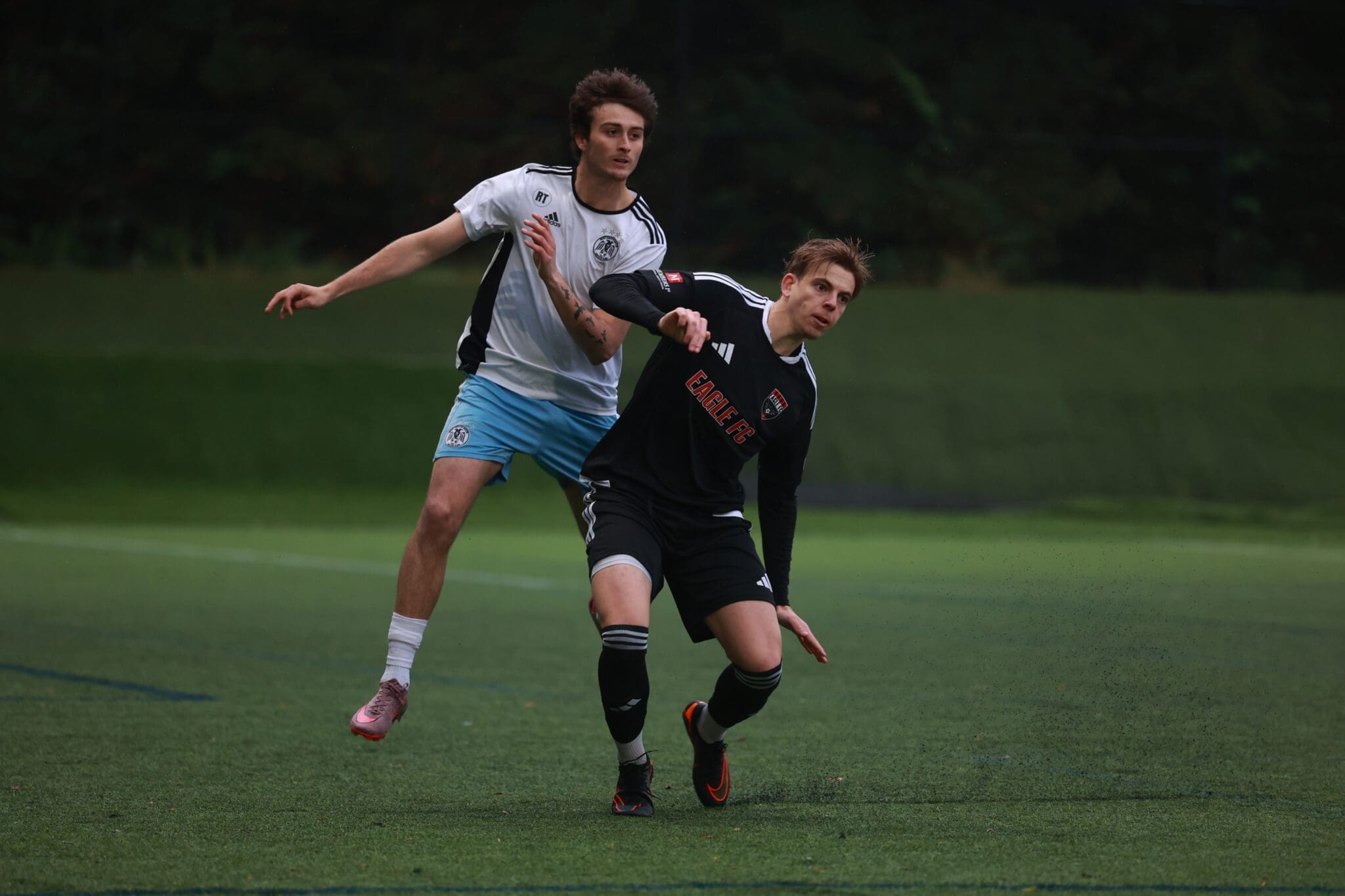 Two soccer players chase the ball on a green field, one in a white jersey and light blue shorts and the other in a black kit with red accents.