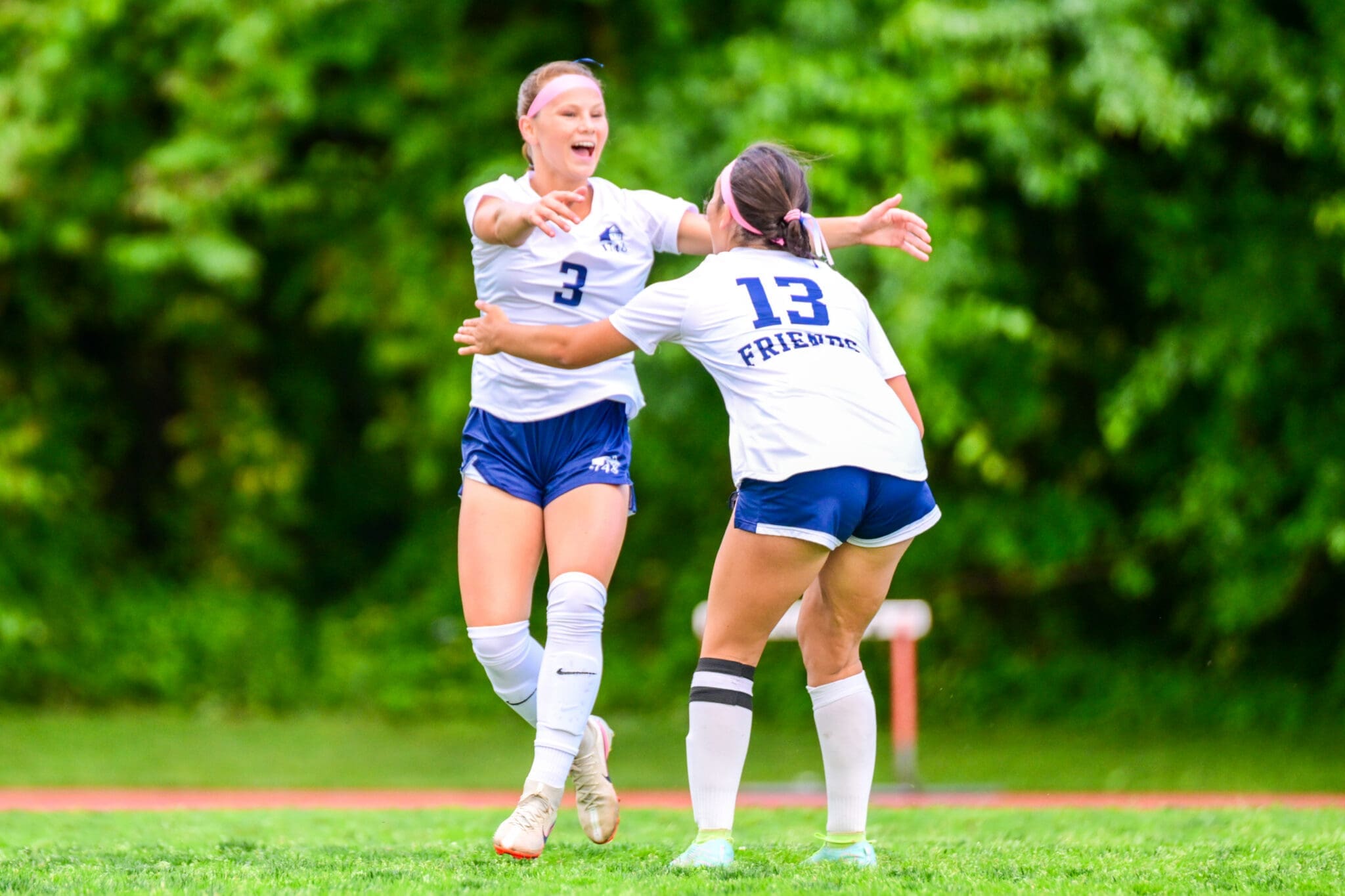 Two female soccer players in white uniforms celebrate together on a green field, arms raised in celebration.