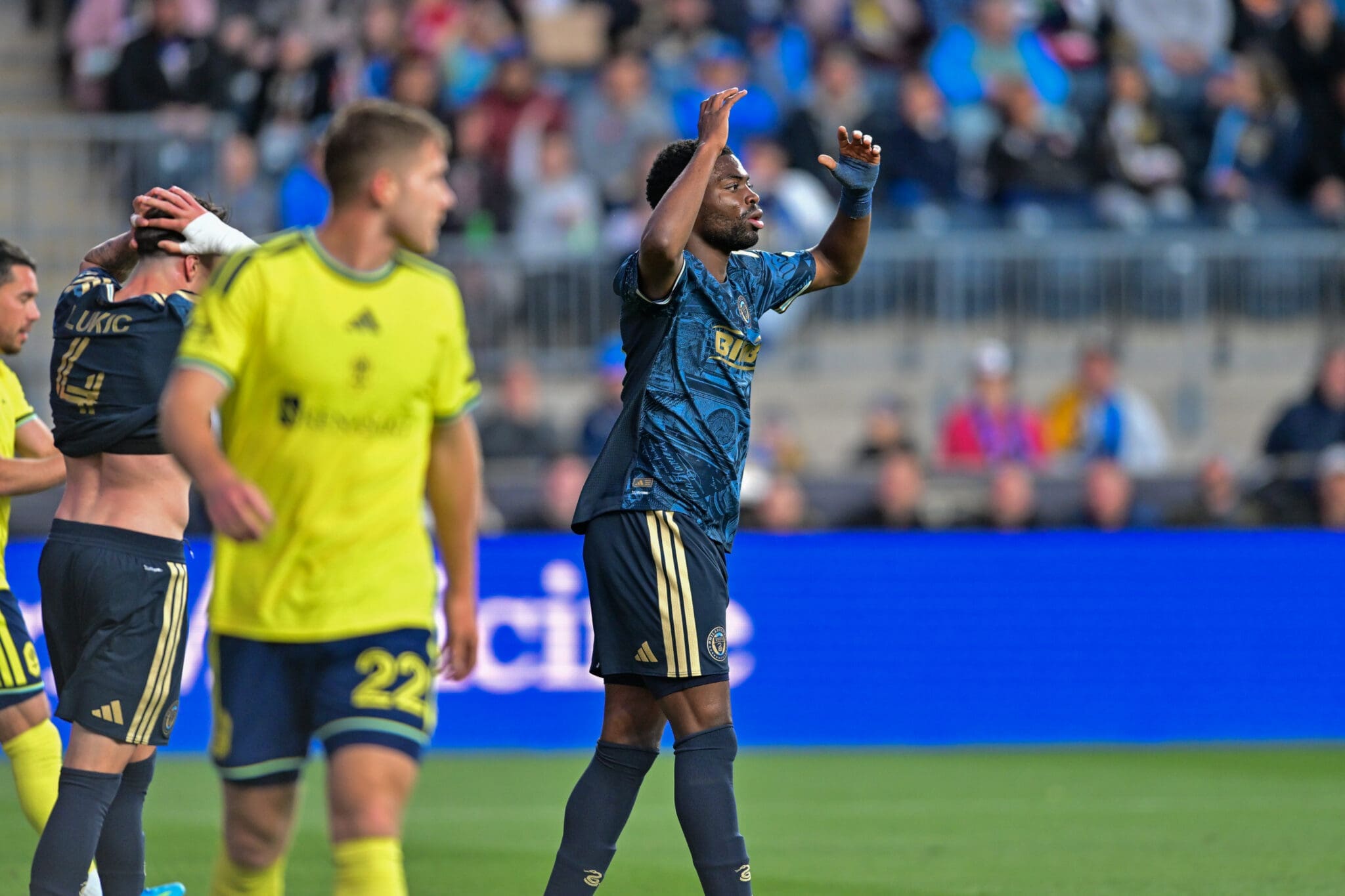 Soccer player in blue raises arms in celebration while teammates in yellow and blue look on on a green field with a blurred crowd in the background.