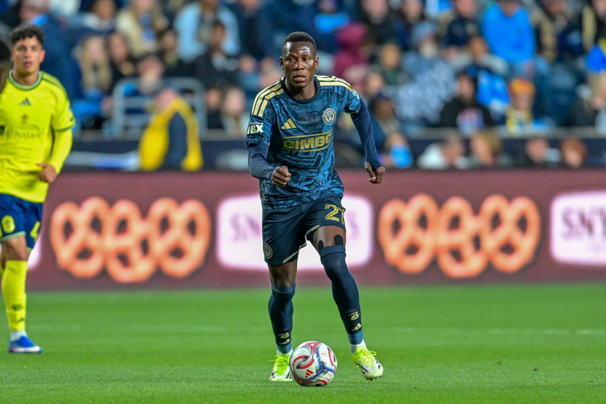 Soccer player in a blue kit dribbles the ball on a green field with a crowd in the stadium behind him, a yellow-clad opponent nearby