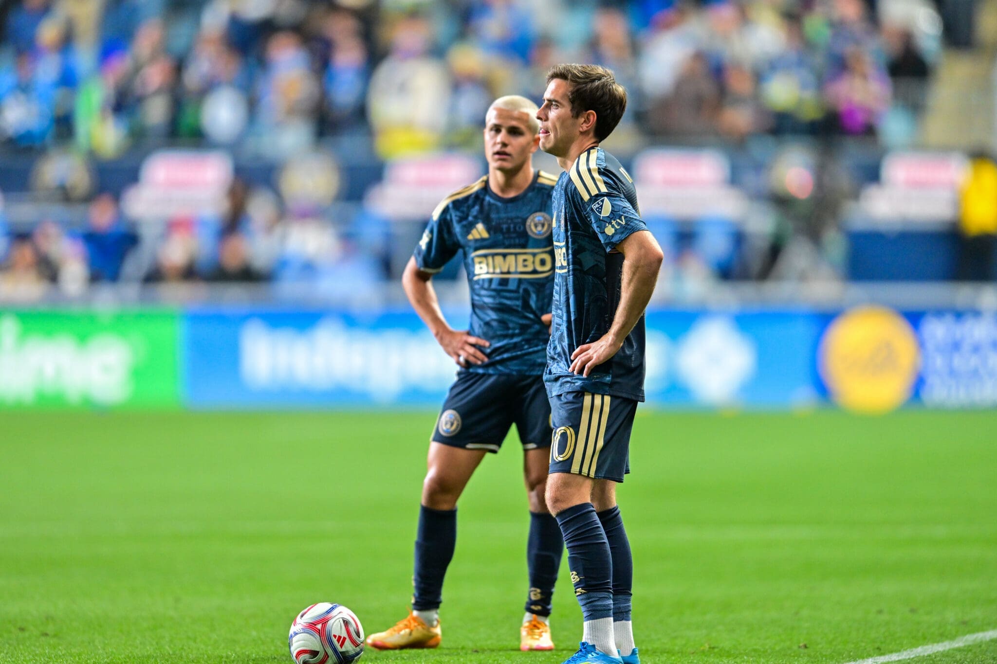 Two soccer players in dark blue uniforms stand on a green pitch with a ball at their feet, blurred stadium crowd behind them.
