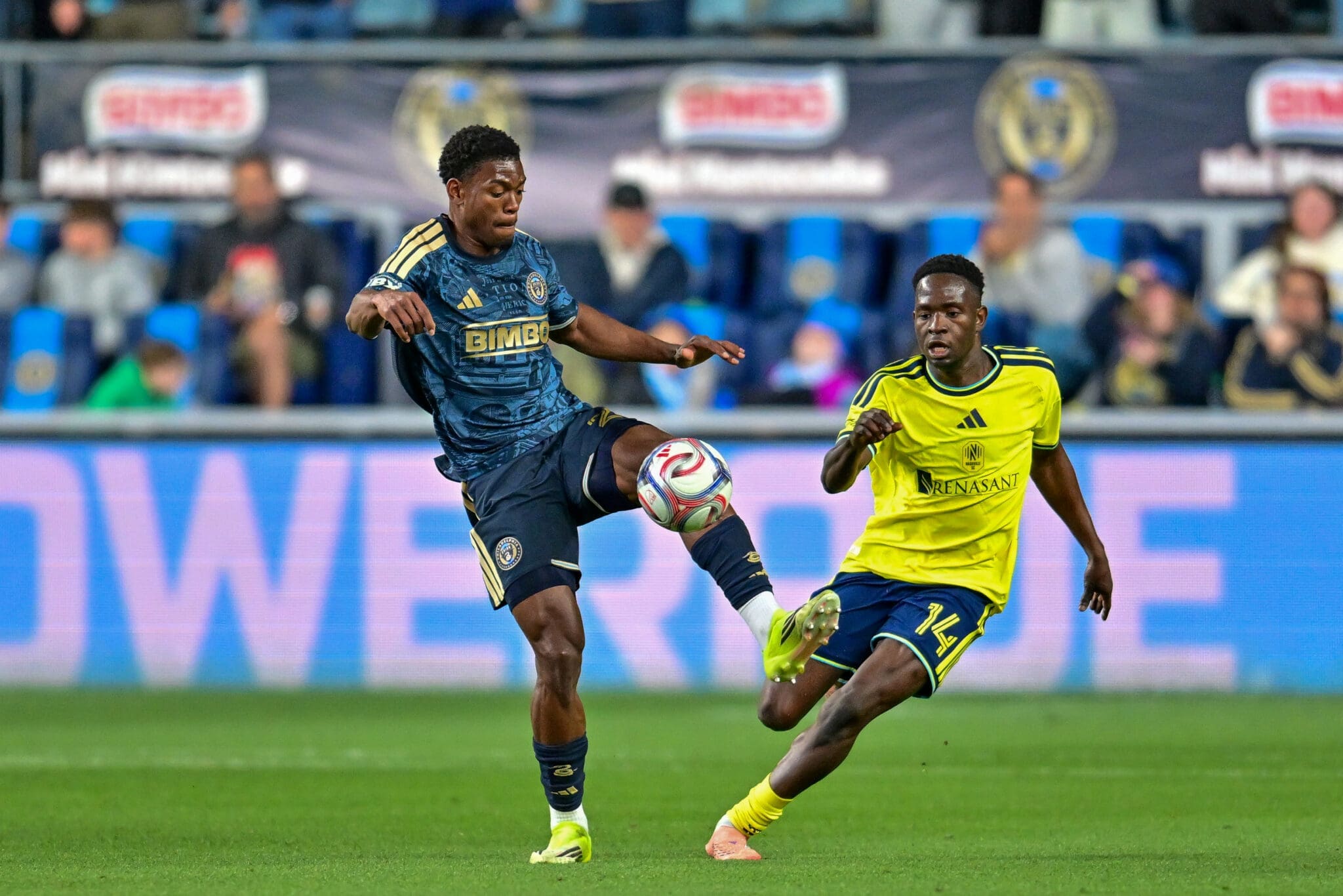 Two soccer players compete for the ball during a match, one in a blue kit and one in a yellow kit, on a green field with spectators in the background.