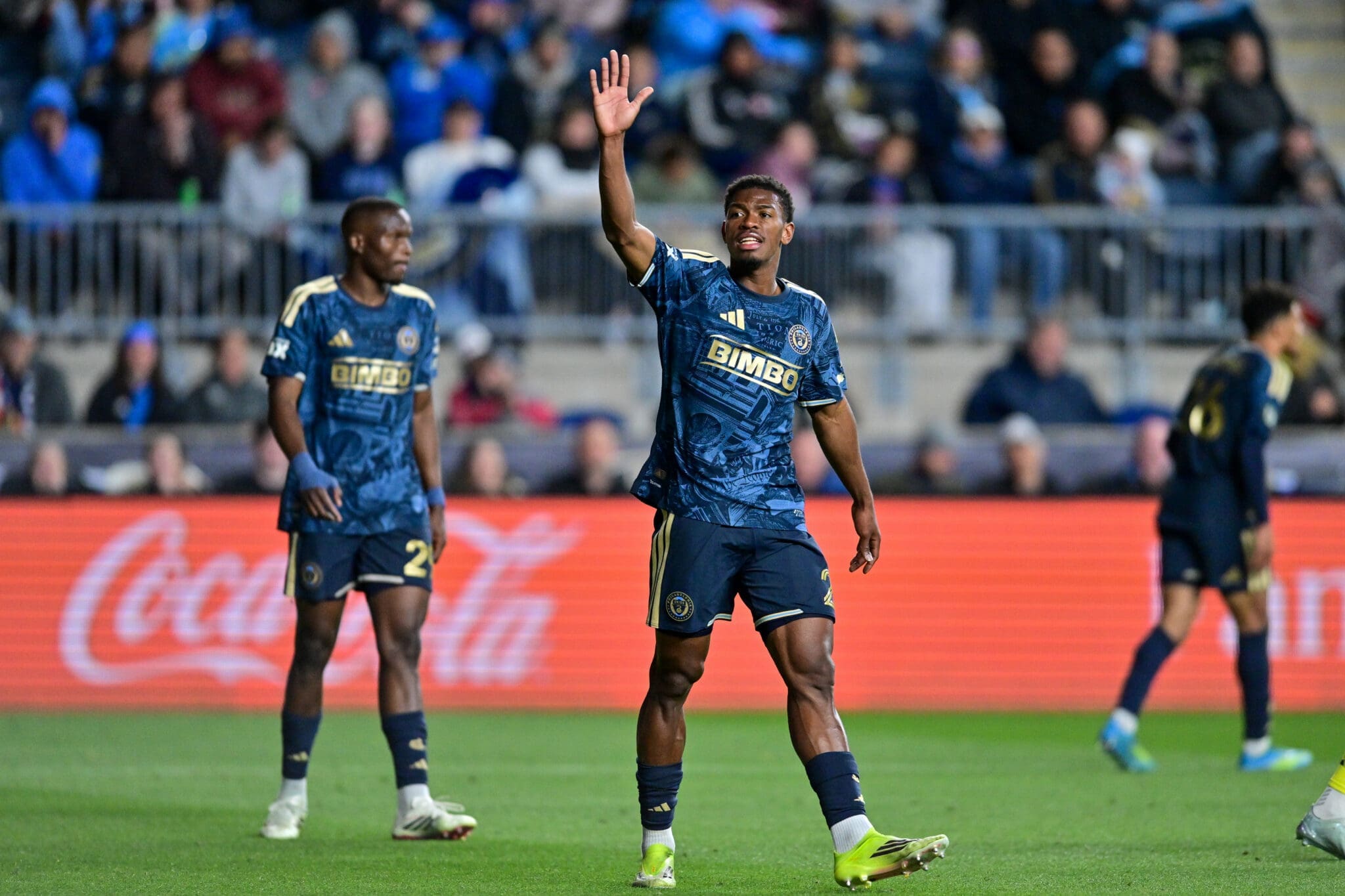 Soccer player in a blue jersey raises his right hand while teammates celebrate on the field.