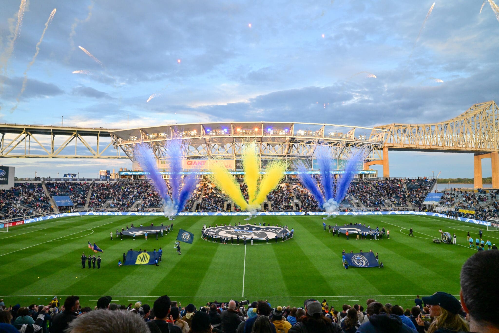 A sports stadium filled with spectators as colorful fountains explode over the field during a ceremony at dusk.
