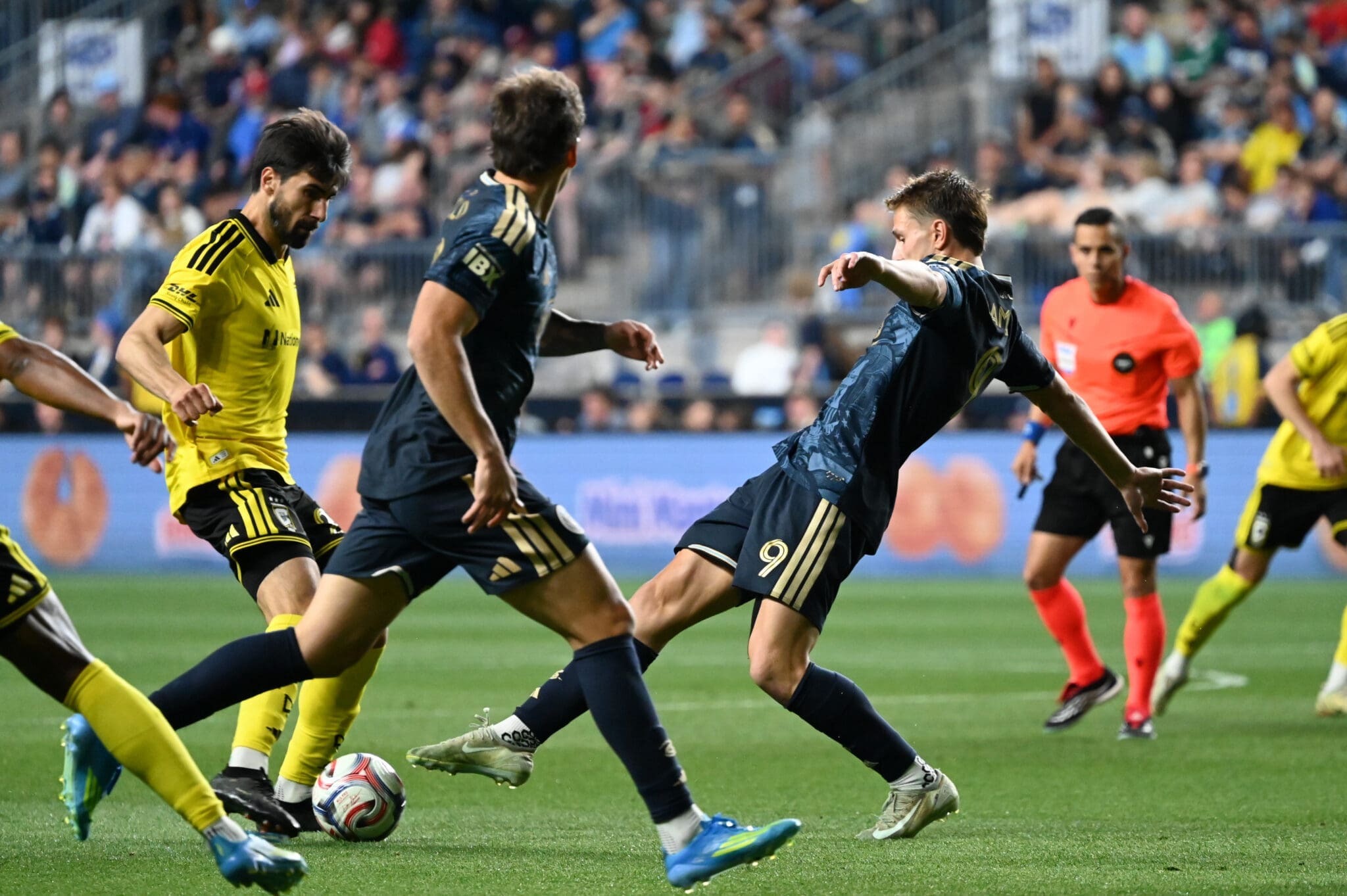 Soccer players in yellow and navy kits chase the ball on a crowded stadium field during a match.