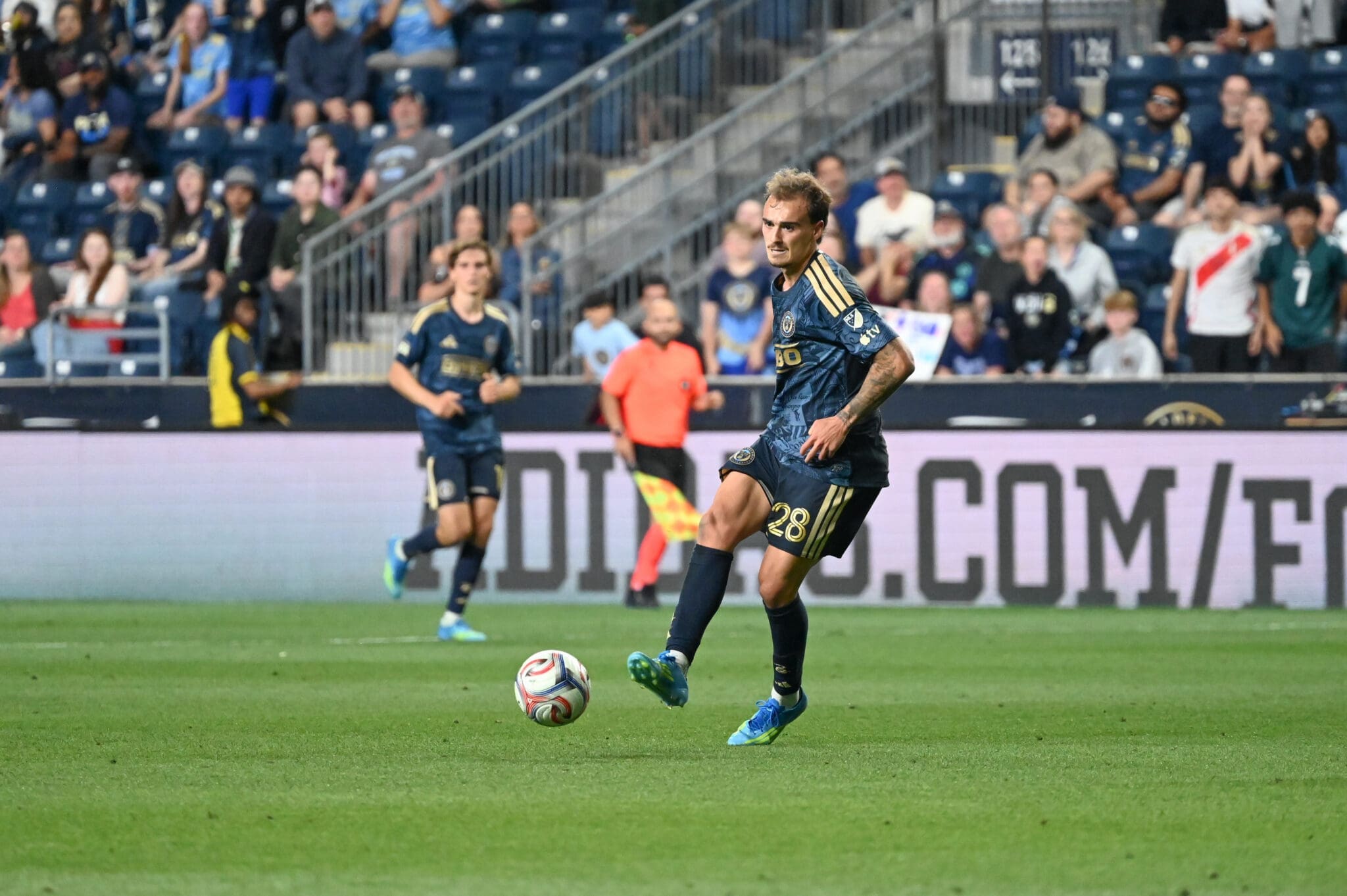 Soccer player in a blue kit with gold stripes (number 28) dribbles a ball on a green pitch as a packed stadium watches from the stands behind him.