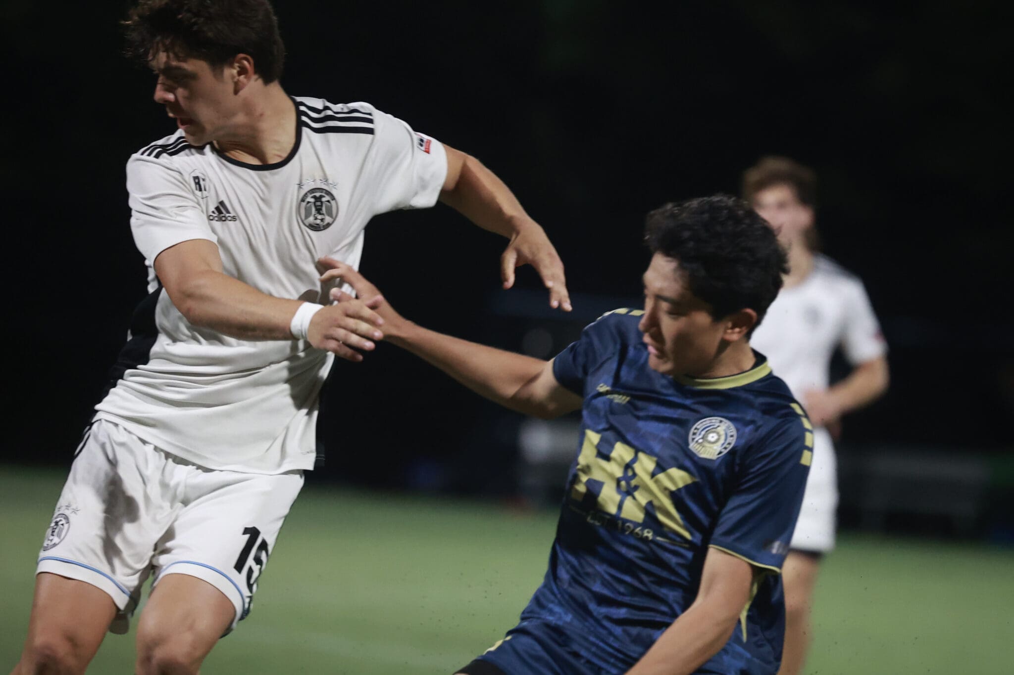 Two soccer players contest the ball on a night field: a white jersey player (#15) shields against a blue-kit opponent.