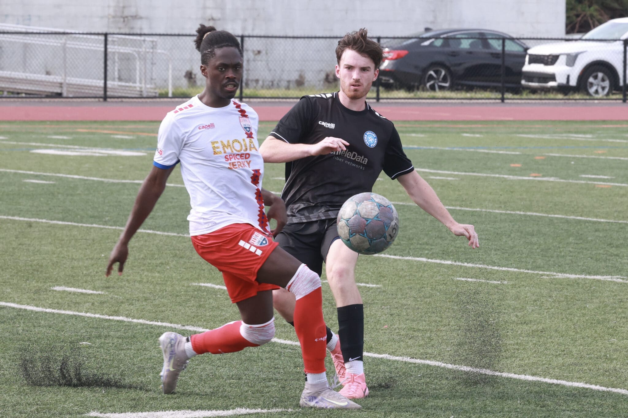 Two soccer players compete for the ball on a grassy field, one in white and red and the other in black, with a ball near their feet and a chain-link fence in the background