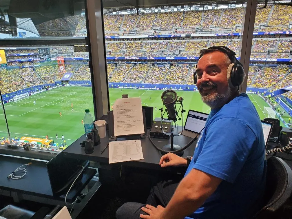 Commentator in a blue shirt wearing headphones, seated at a broadcast desk with microphone, overlooking a packed stadium.